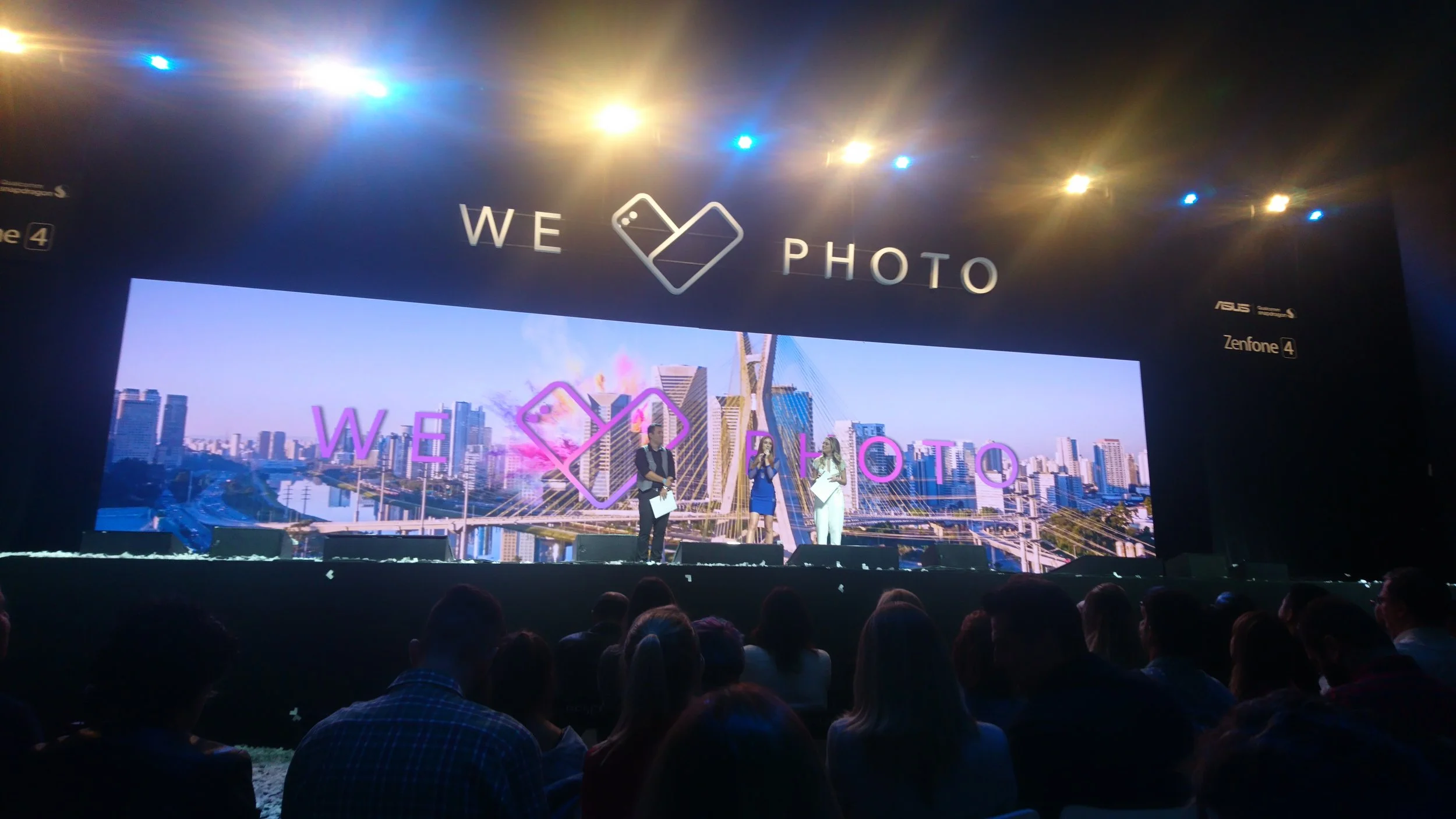 Stage with large screen displaying city skyline and pink-hearts graphic, with three people speaking into microphones, audience seated in front.
