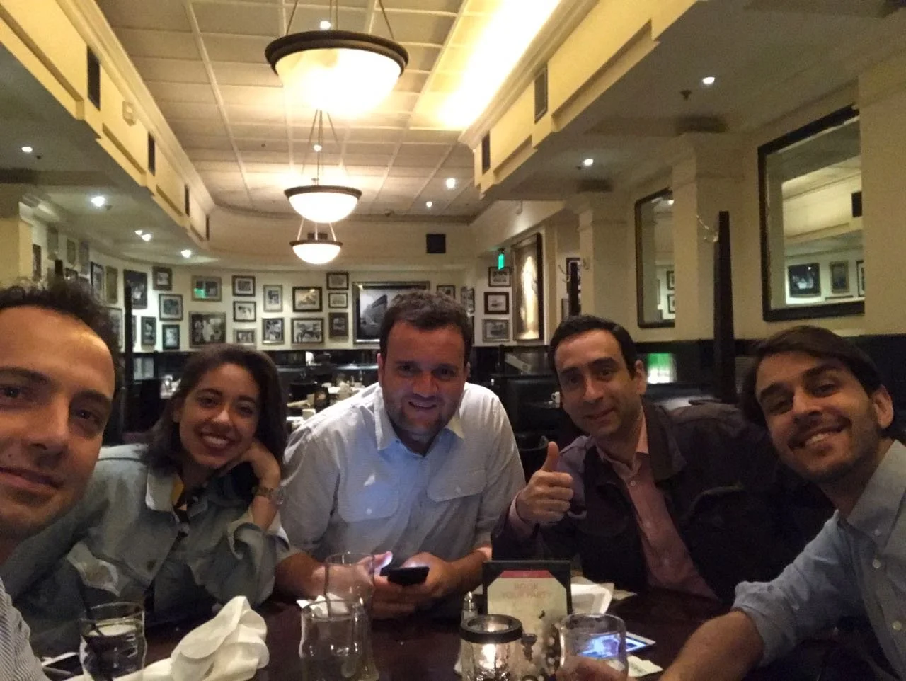 Five friends sitting at a restaurant table, smiling for the camera. The background features framed pictures on the walls and warm lighting.