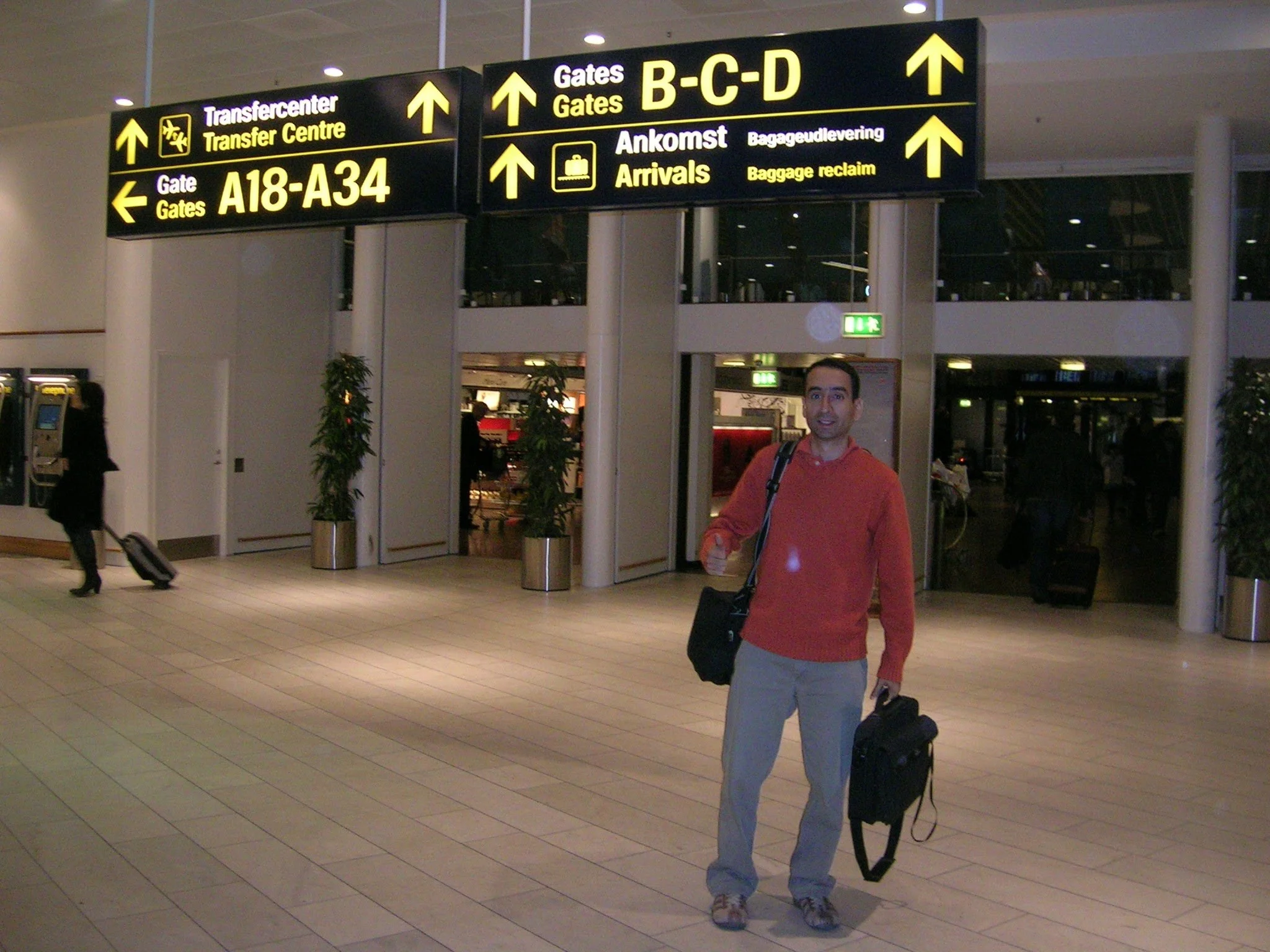 A man standing inside an airport terminal, holding a small rolling suitcase and a shoulder bag, with airport signage above him indicating directions to gates, arrivals, and baggage reclaim.