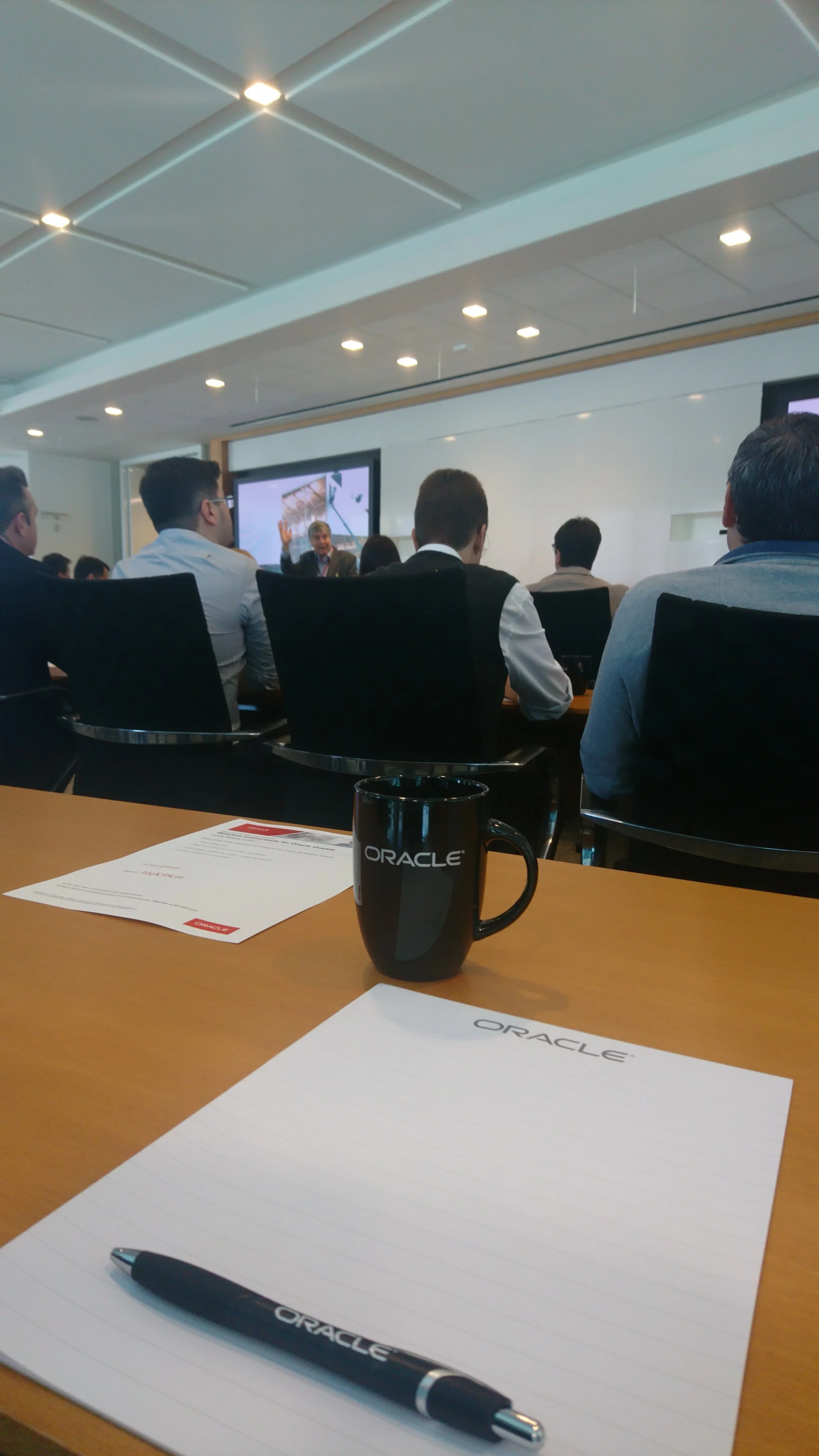 A conference room with people attending a presentation, a black Oracle coffee mug, lined notepad with Oracle logo, and a pen on a wooden table.