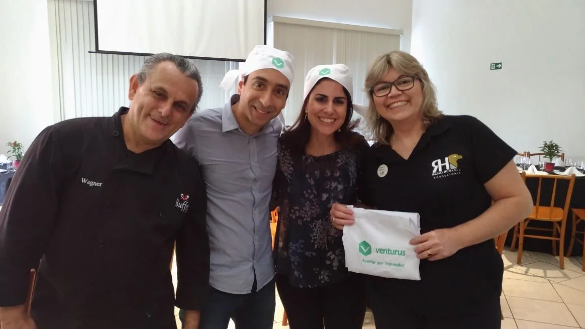 Four people smiling at the camera, standing together in a restaurant or event space. Two women are wearing white hats with a green check mark, and one woman is holding a white bag with the Venturus logo. The men are dressed casually, one in a chef's 