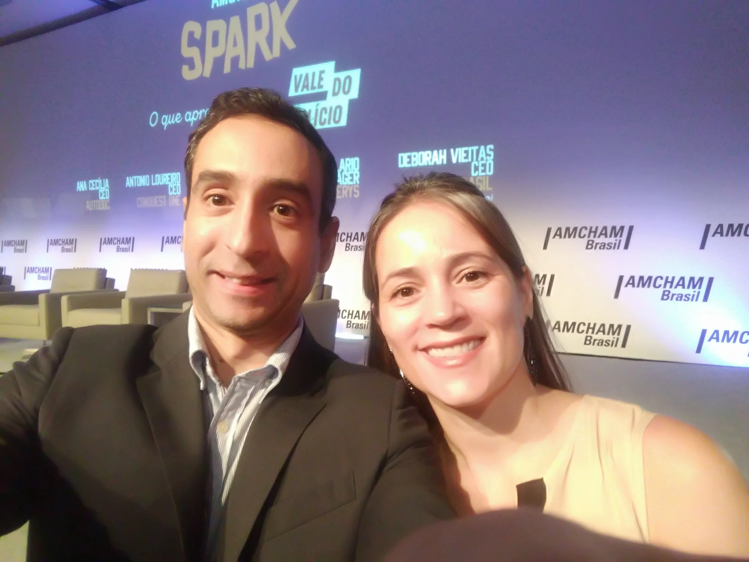 Two people taking a selfie at a conference, standing in front of a backdrop with the logo 'AMCHAM Brasil' and various names. The man on the left is wearing a black suit and striped shirt, and the woman on the right is wearing a light-colored sleevele