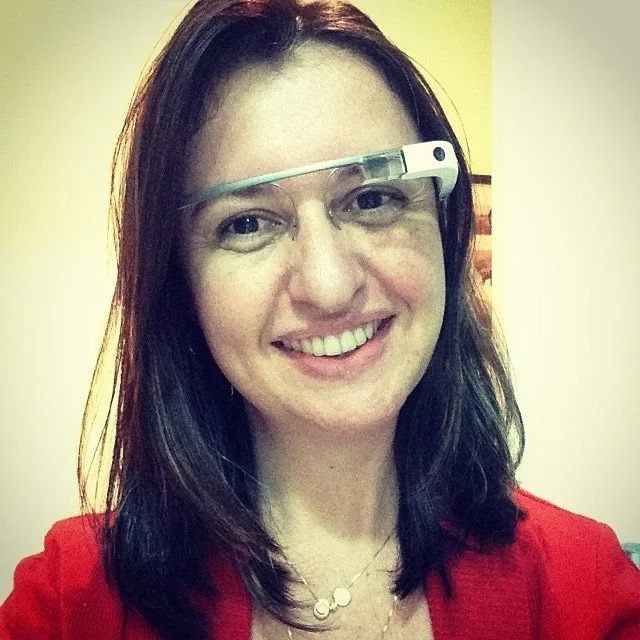 A woman with shoulder-length dark hair wearing Google Glass and a red top, smiling indoors.