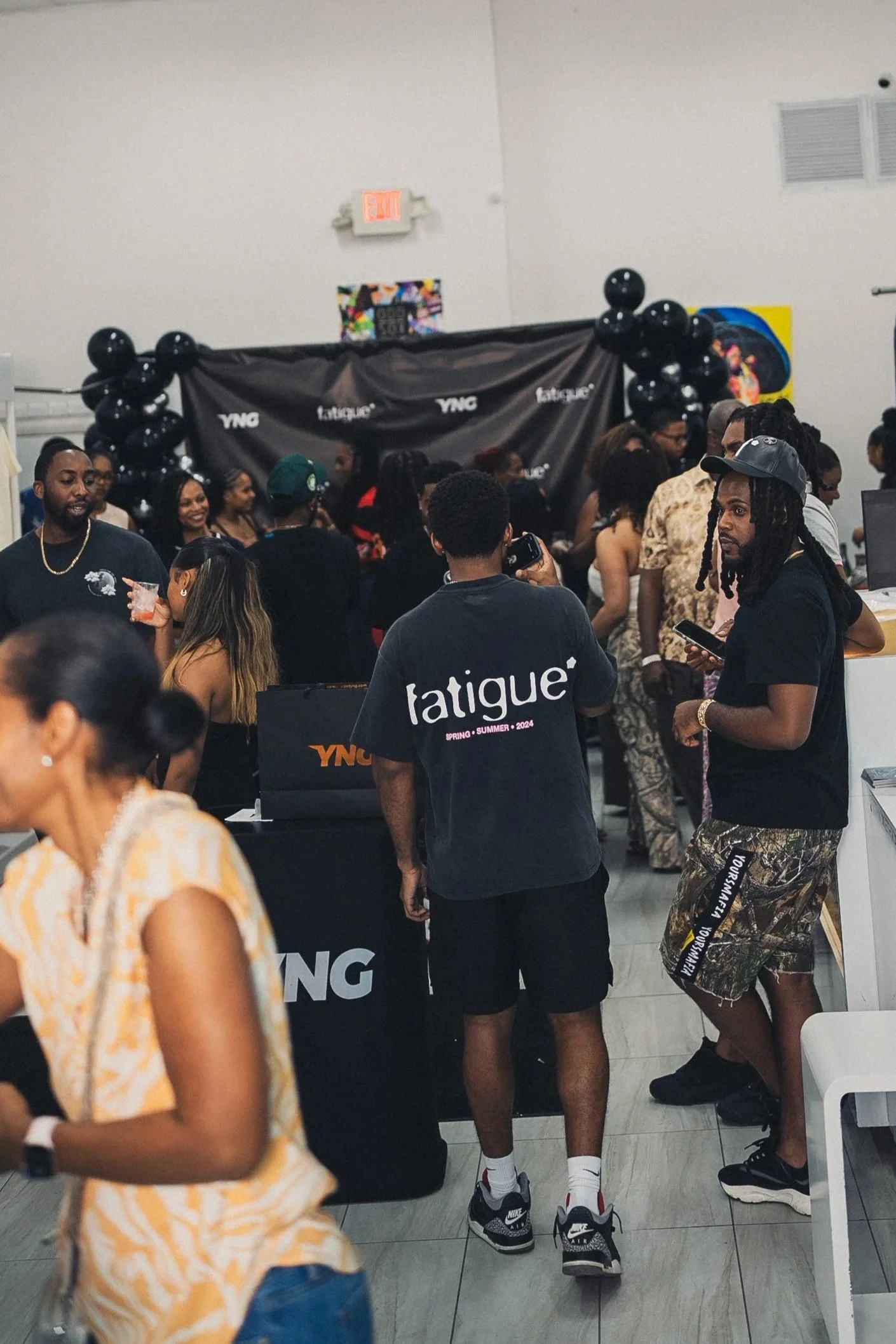 A crowded indoor event with people mingling, some holding drinks, and a backdrop with balloons and branding for 'YNG' and 'tatique'.