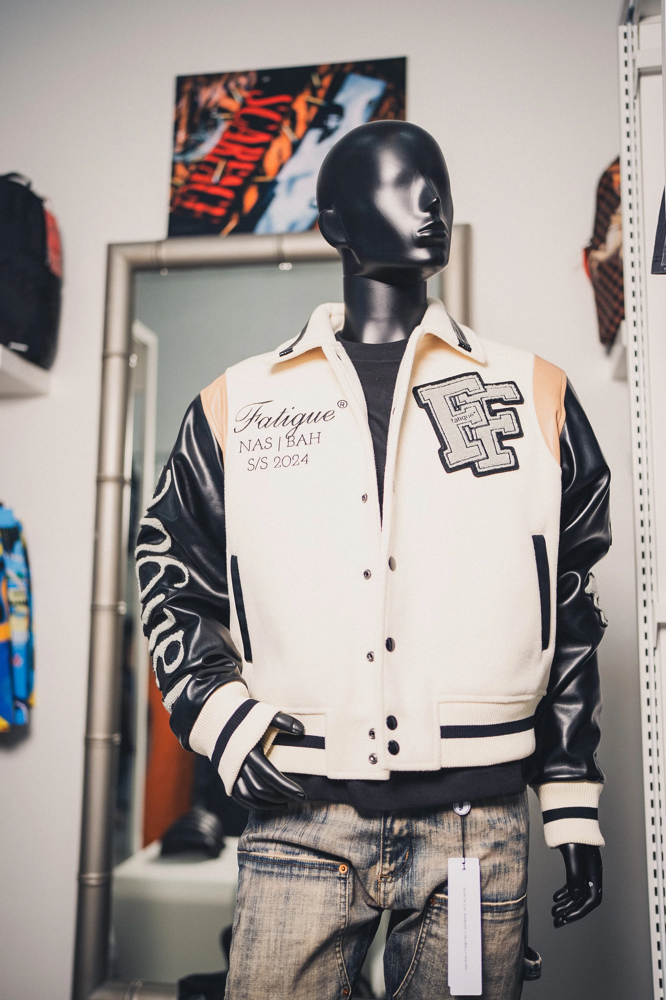 A black mannequin wearing a varsity jacket with black leather sleeves and cream body, with patches and embroidery, paired with distressed jeans, displayed in a retail store.