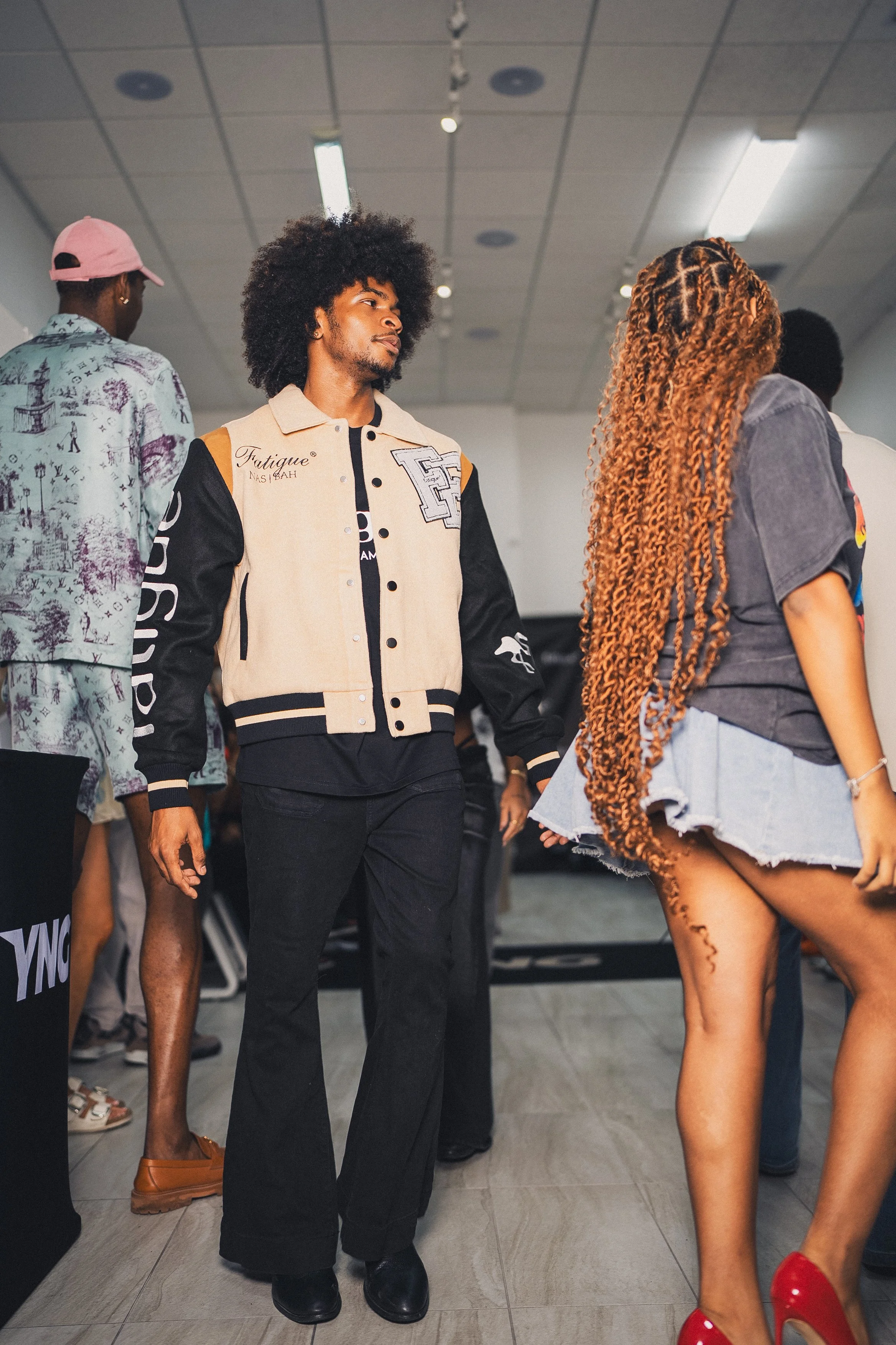 People walking in an indoor space, possibly a store or event. The man in the foreground has curly hair, wears a beige and black varsity jacket, and black pants. A woman with long, bundled hair in a gray T-shirt and denim shorts is nearby. These are c