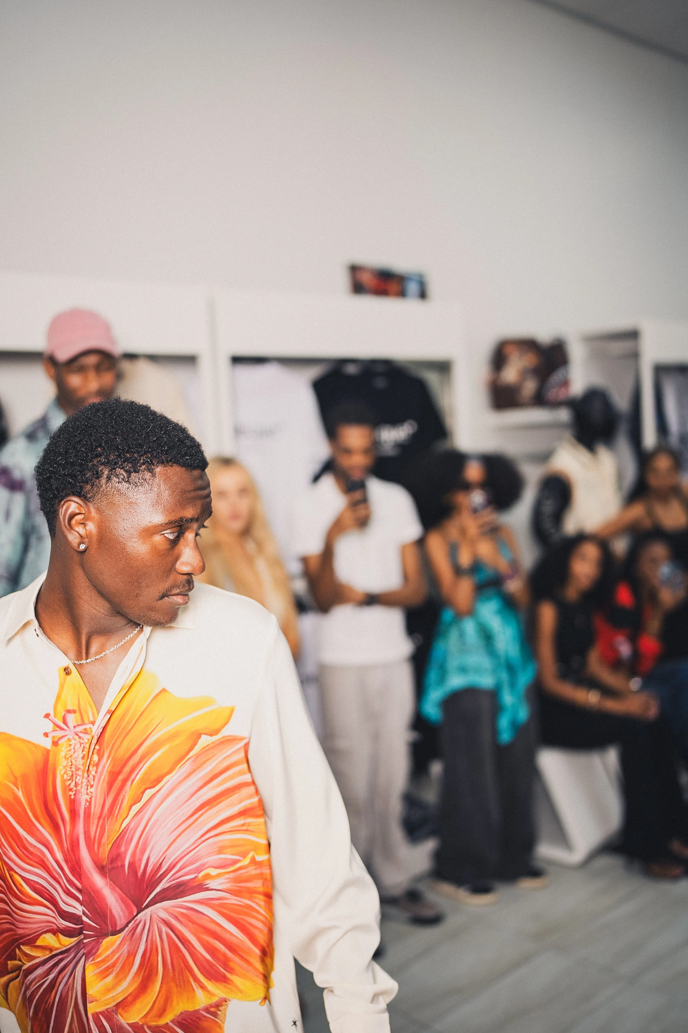 A man in a shirt with a large orange and yellow flower design on the front, standing in a room filled with people, some of whom are watching or taking photos.