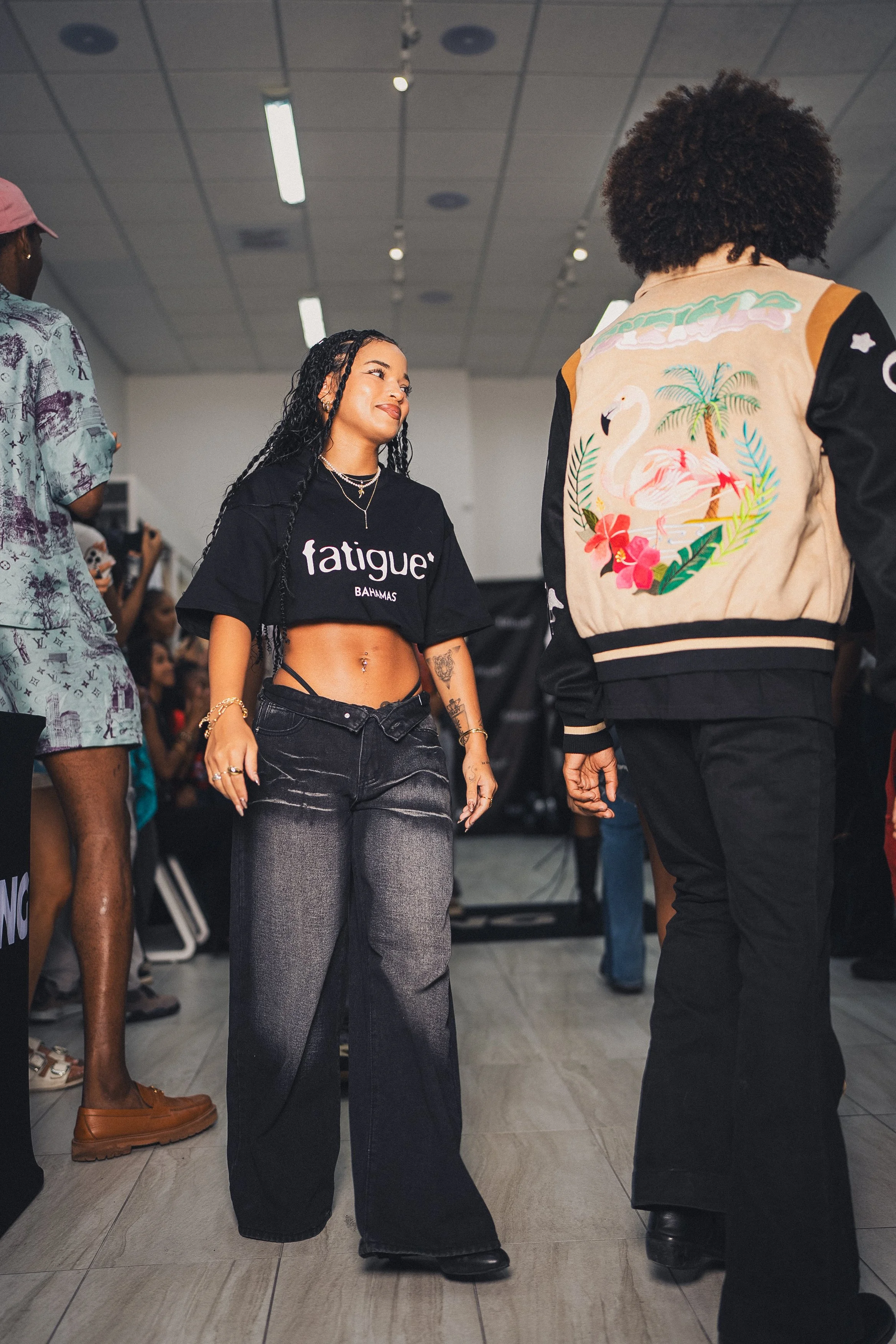 A young woman with long braids, wearing a black crop top that says 'fatigue BAYAMAS', black wide-leg jeans, and jewelry, is looking at a person with an afro hairstyle and a jacket with a tropical flamingo design, in a crowded indoor space.