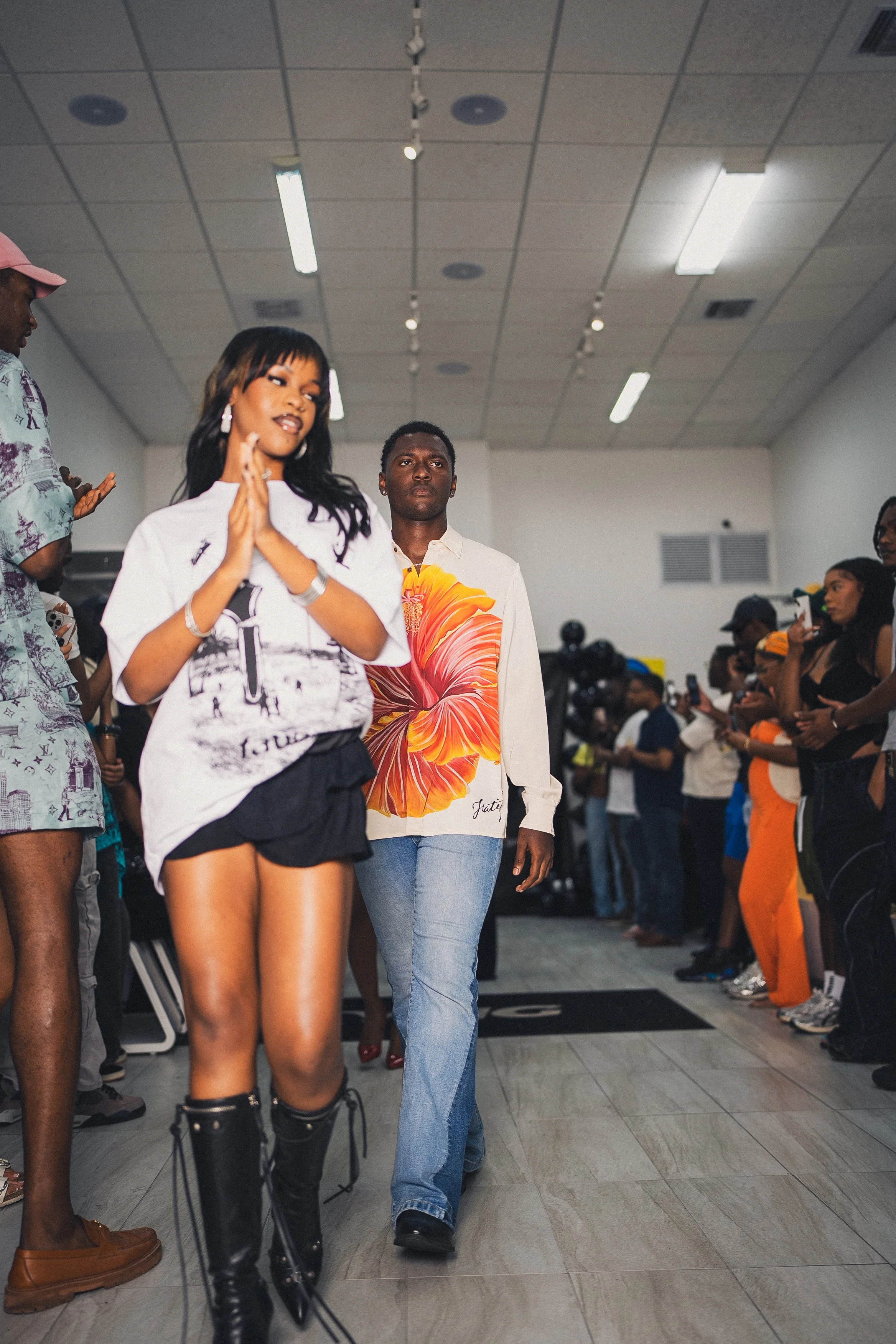Two models walking down a runway at a fashion show, surrounded by seated and standing spectators taking pictures. The female model in front is wearing a white t-shirt with a black and white graphic, black shorts, and tall black boots. The male model