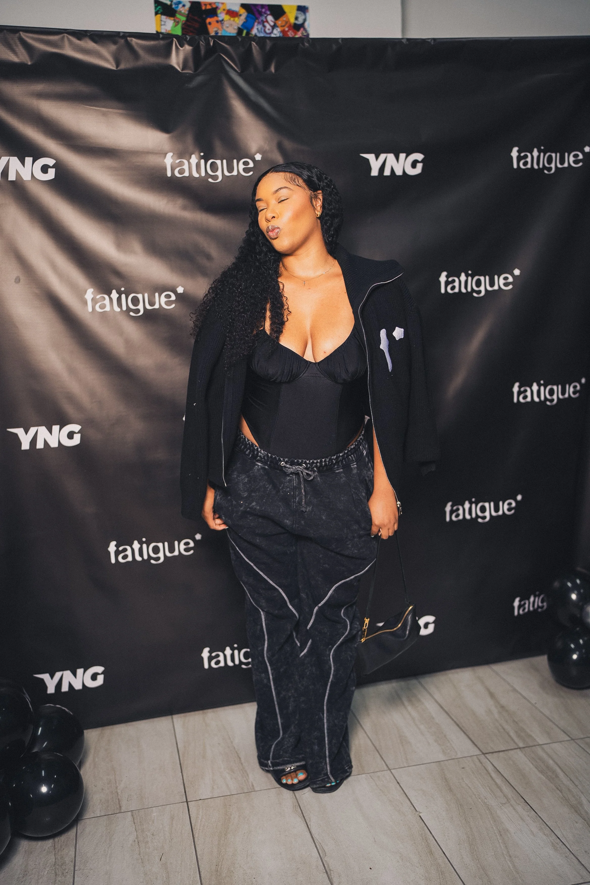A woman with long curly black hair standing in front of a black backdrop featuring the words 'YNG' and 'fatigue'. She is wearing a black top, black pants with white seams, a black zip-up jacket draped over her shoulders, and black heels, posing with