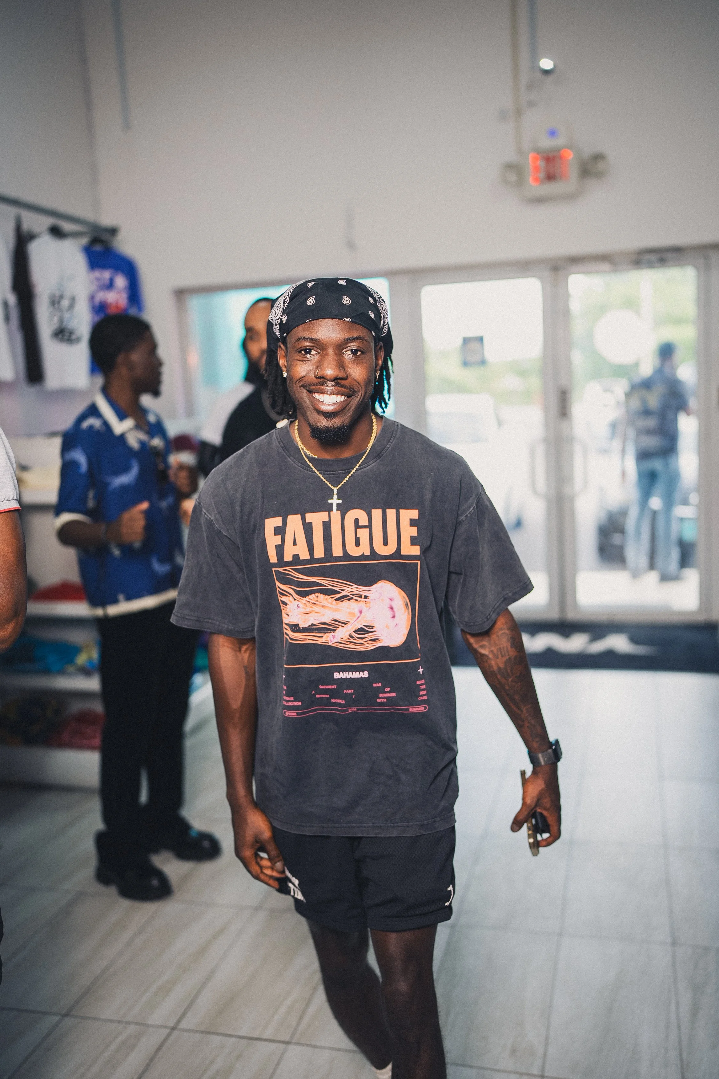 A smiling man walking inside a store, wearing a black t-shirt with 'FATIGUE' in large letters and a graphic of a jellyfish, black shorts, a bandana, and a gold chain.