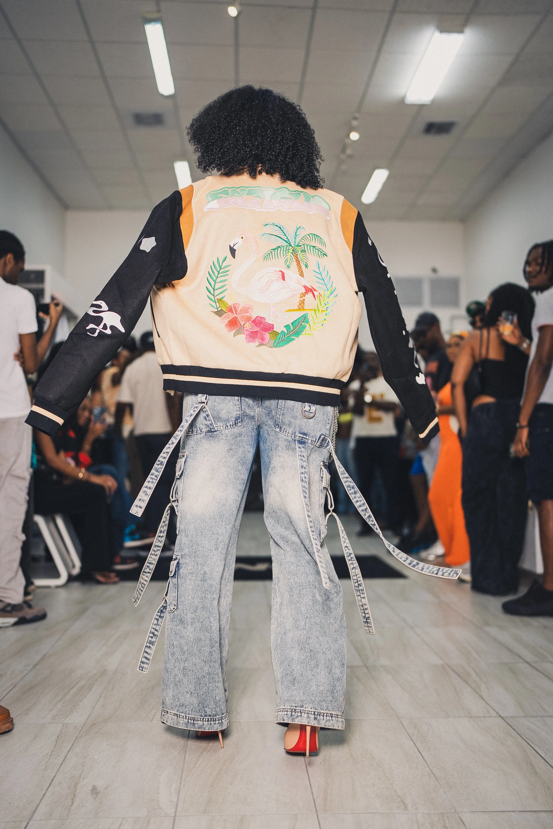 Person modeling a jacket with a flamingo and tropical scene, wearing wide-leg jeans with hanging straps, in a crowded indoor space.