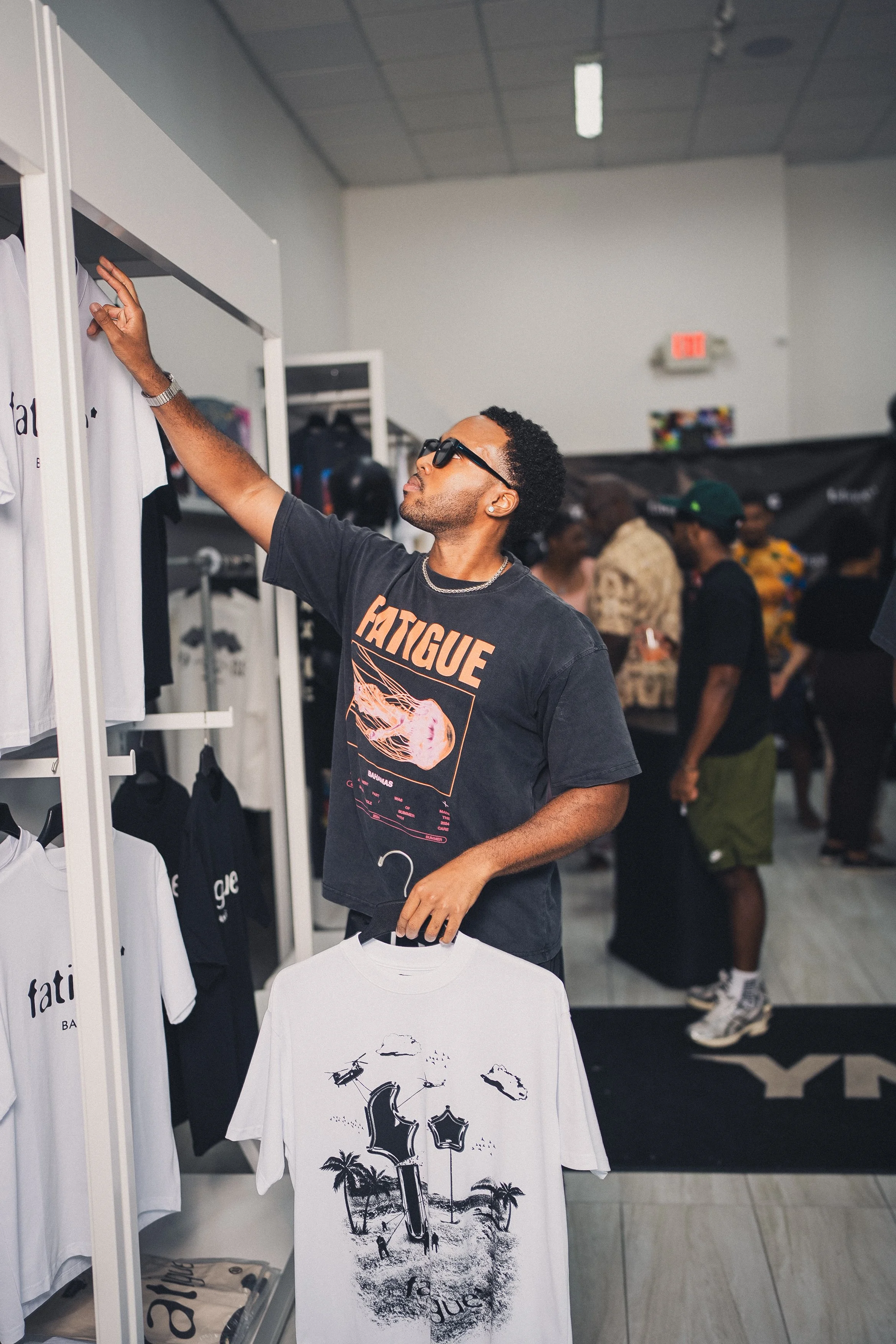 A man wearing sunglasses and a black T-shirt with the word "FATIQUE" on it is shopping for T-shirts at a clothing store, holding a white T-shirt with a graphic print of palm trees and a figure on it.