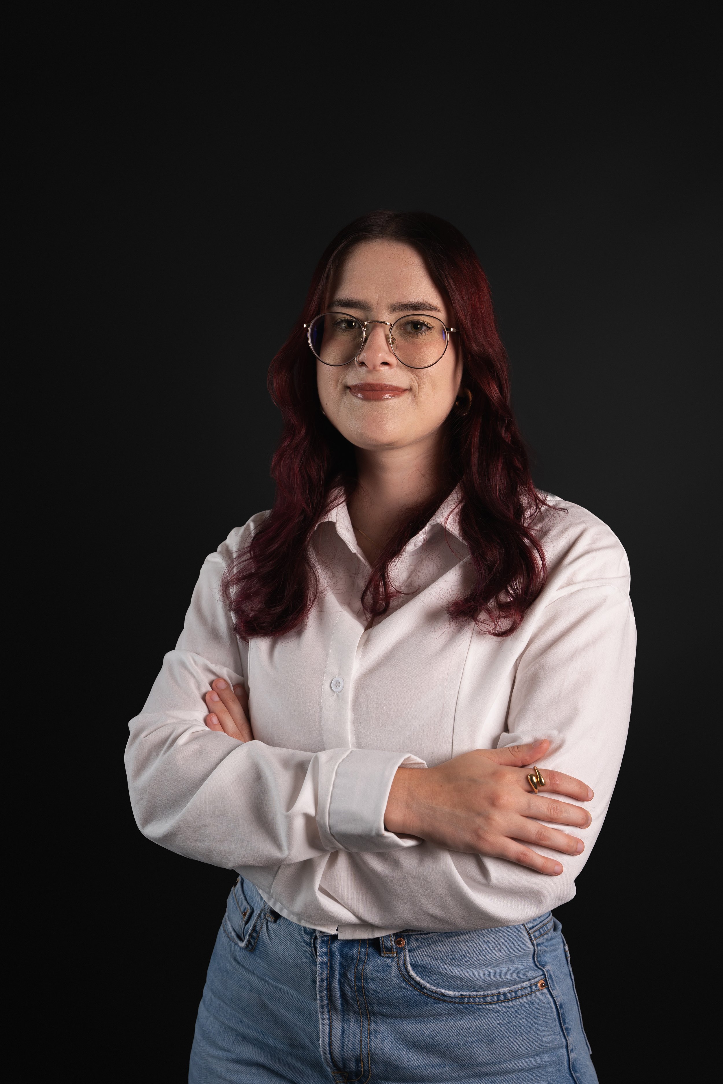 Person with long hair and glasses wearing a white shirt and blue jeans standing against a black background with arms crossed.