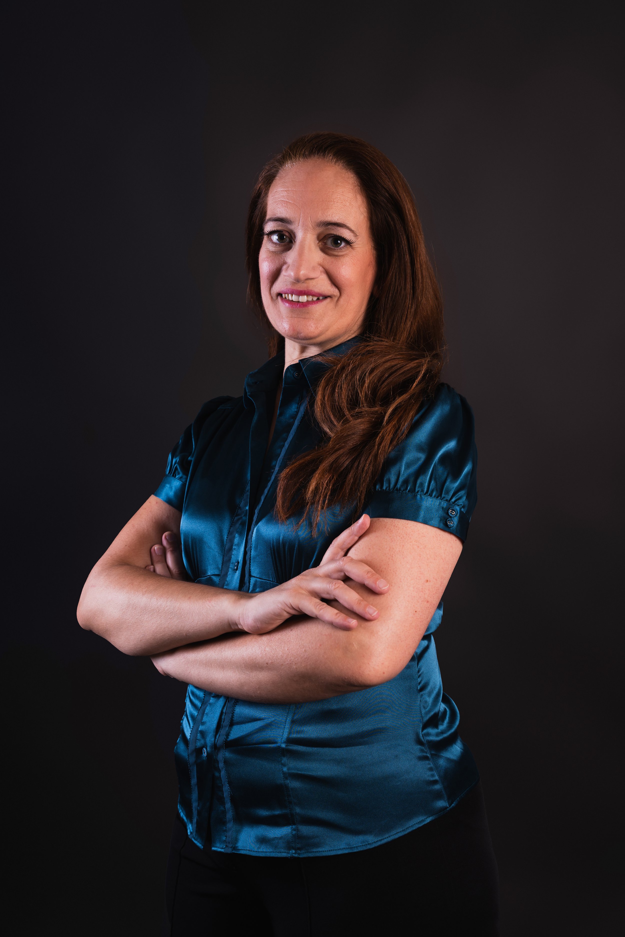Woman in blue blouse with arms crossed against dark background.
