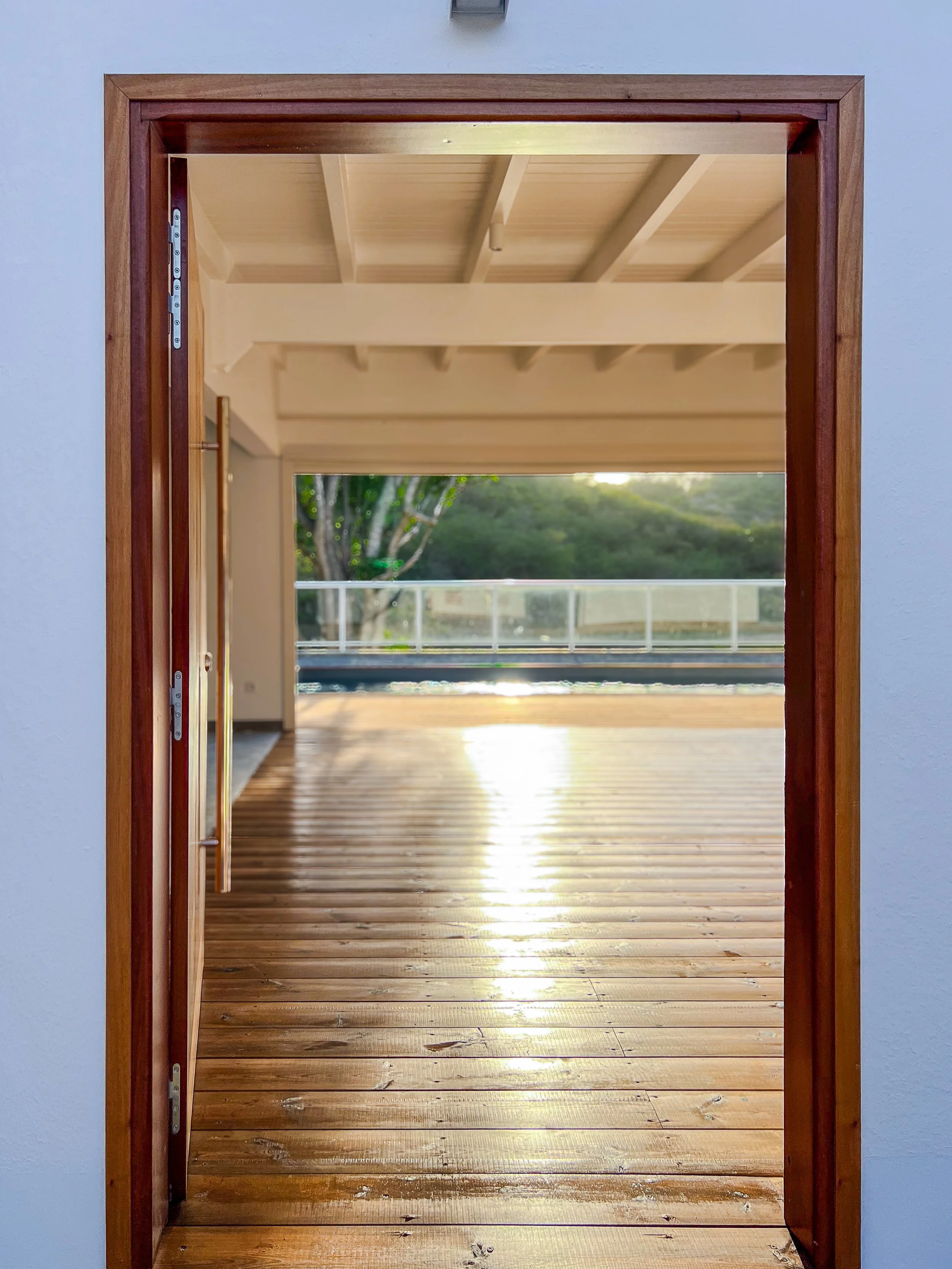 Une porte en bois s'ouvre sur une terrasse en bois avec vue sur un espace extérieur avec des arbres et une barrière en verre, sous un plafond en bois. Le soleil se reflète sur le sol.