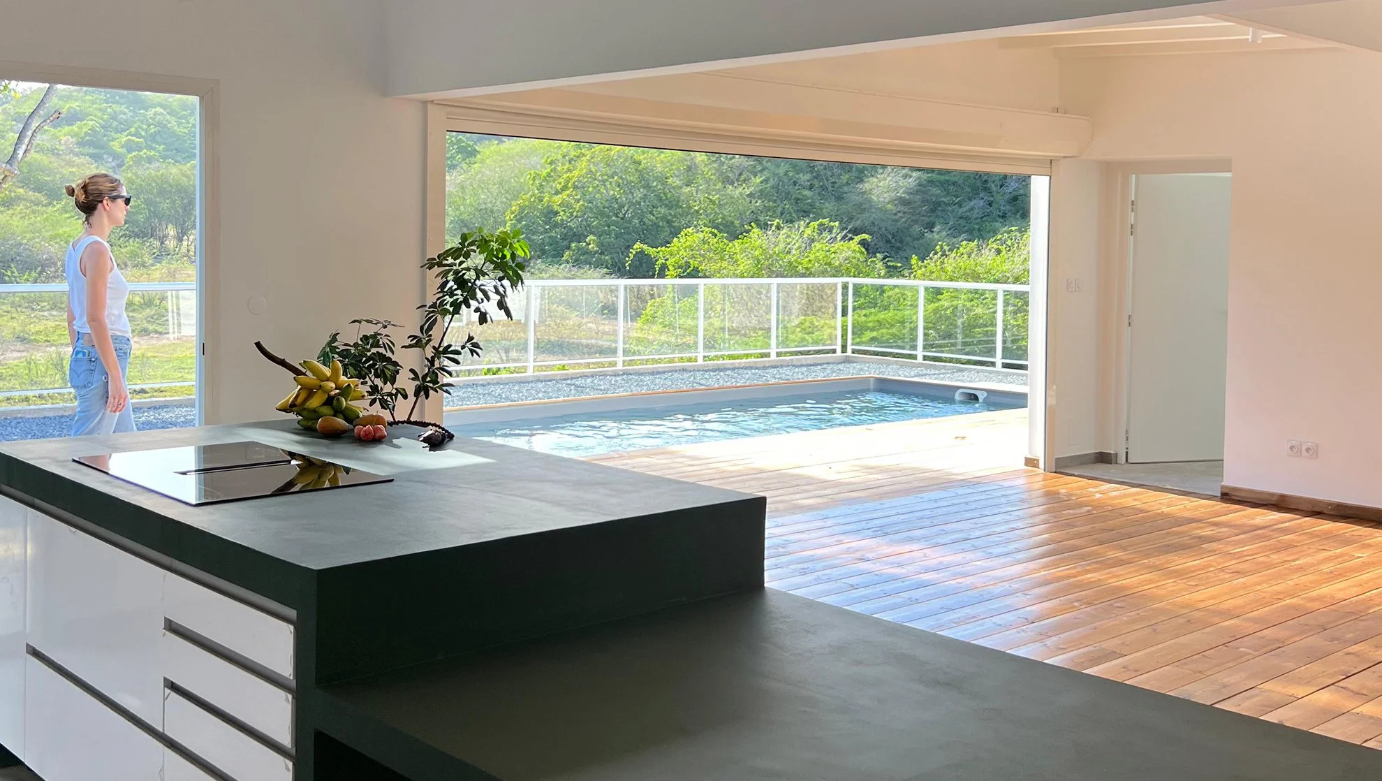 Intérieur d'une maison moderne avec une cuisine ouverte donnant sur une piscine et un espace extérieur avec des arbres verdoyants, un homme avec des lunettes de soleil et un t-shirt blanc, vue depuis la cuisine