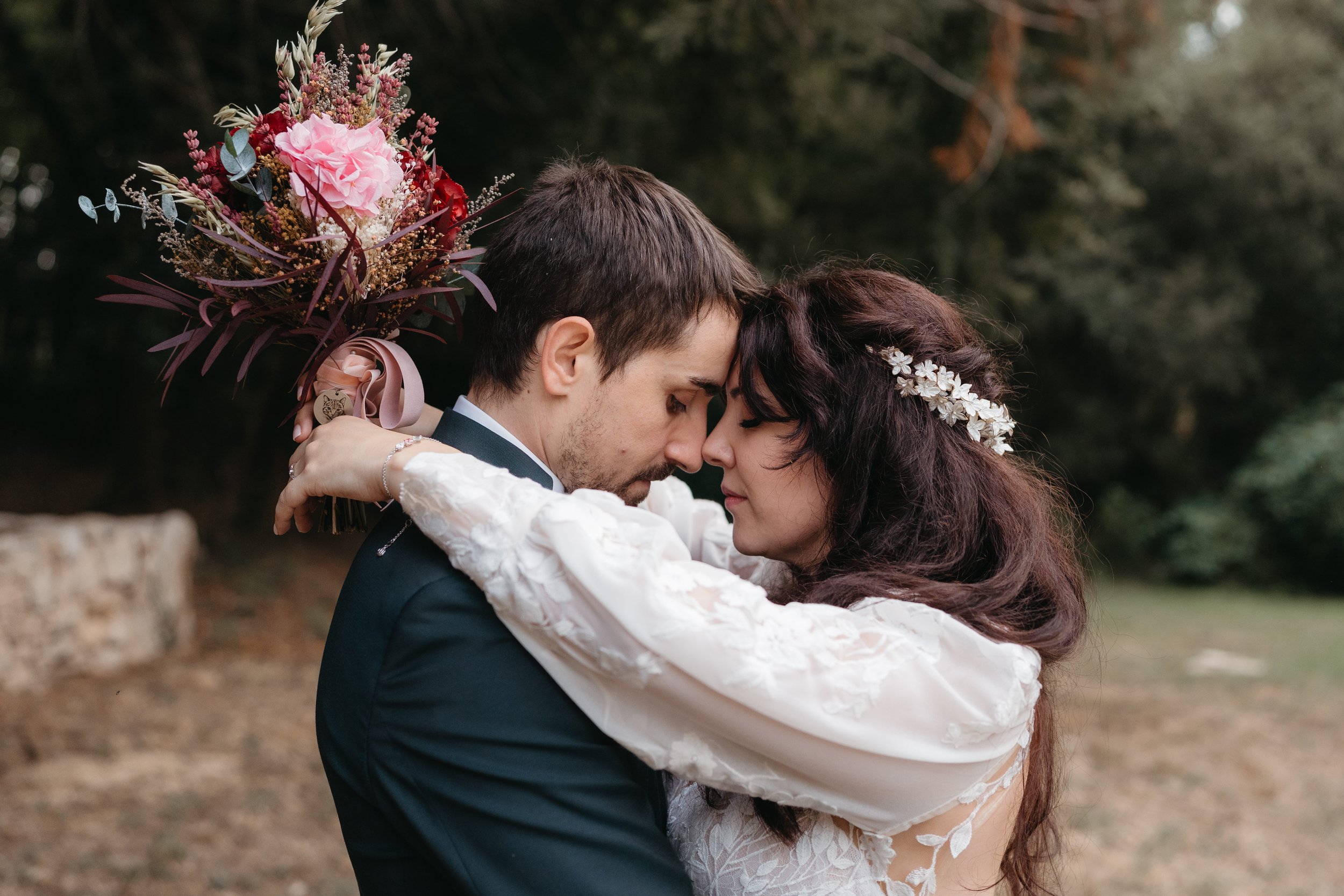 Una pareja de novios en un abrazo con la frente tocándose, en un entorno natural, ella lleva un vestido de novia y una corona de flores, él viste un traje formal, ella sostiene un ramo de flores en la espalda.