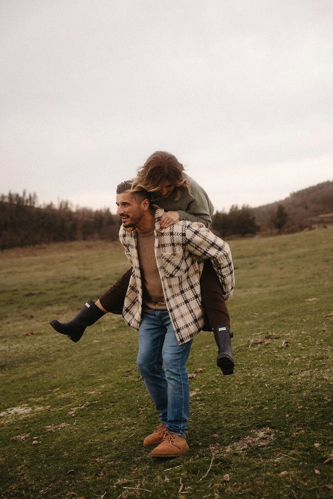 Un hombre lleva en la espalda a una mujer en un campo verde cercano a colinas. Ambos parecen felices y están vestidos con ropa de abrigo. Es un día nublado.
