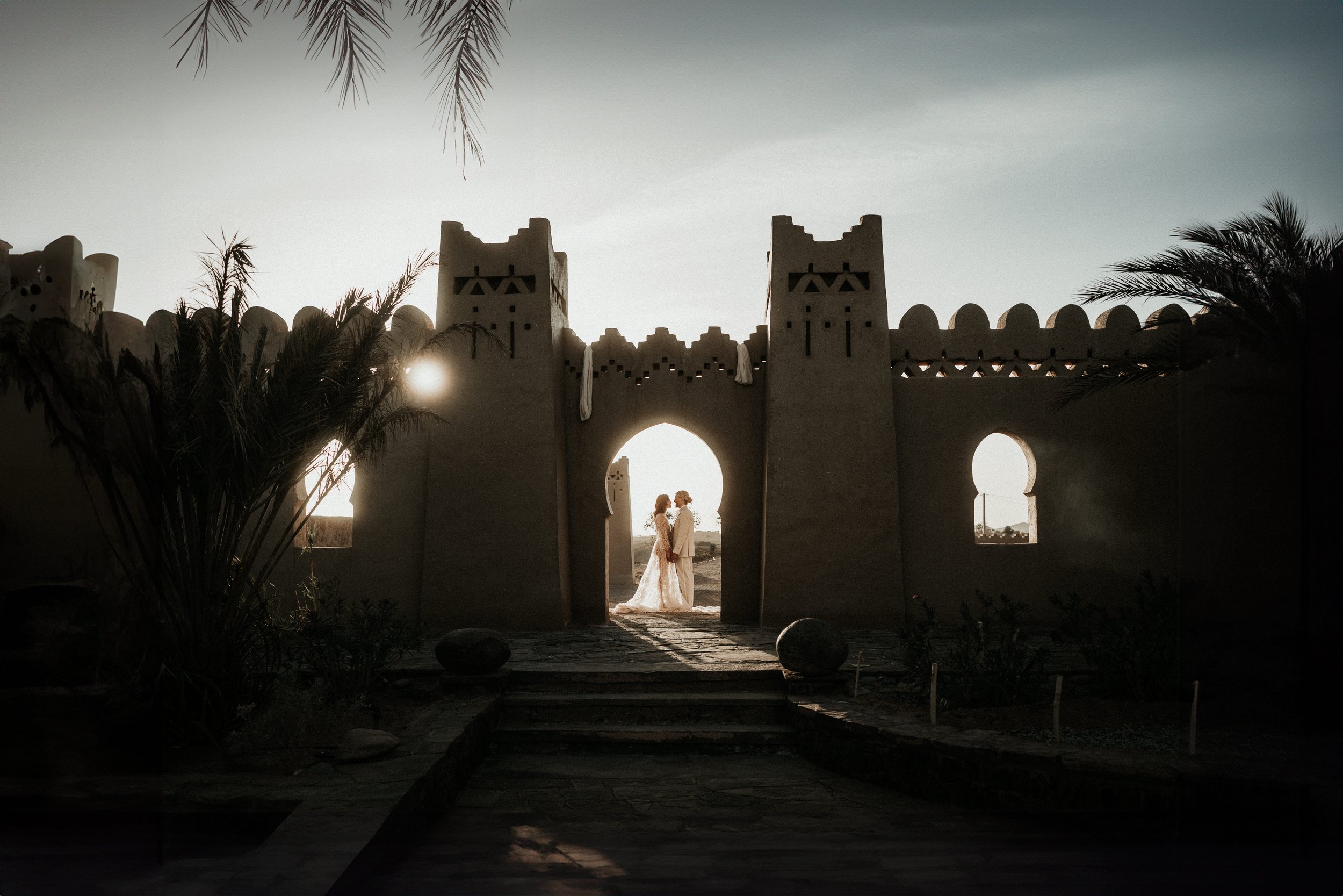 Pareja de recién casados en la entrada de una estructura arquitectónica tradicional, en un ambiente con luz suave y plantas alrededor.