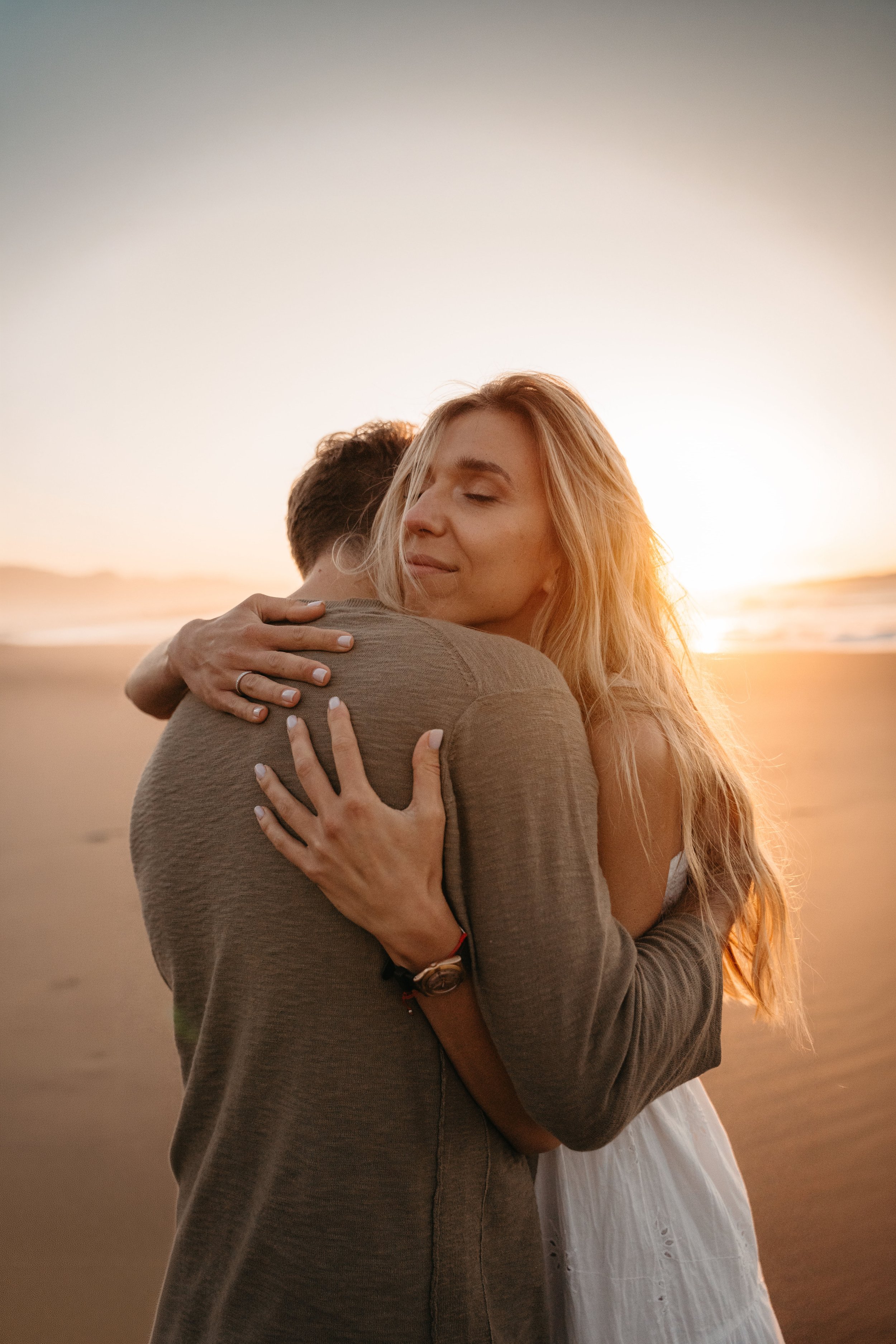 Una pareja abrazándose en la playa al atardecer, la mujer con cabello rubio y expresión de felicidad, el hombre con cabello castaño oscuro.