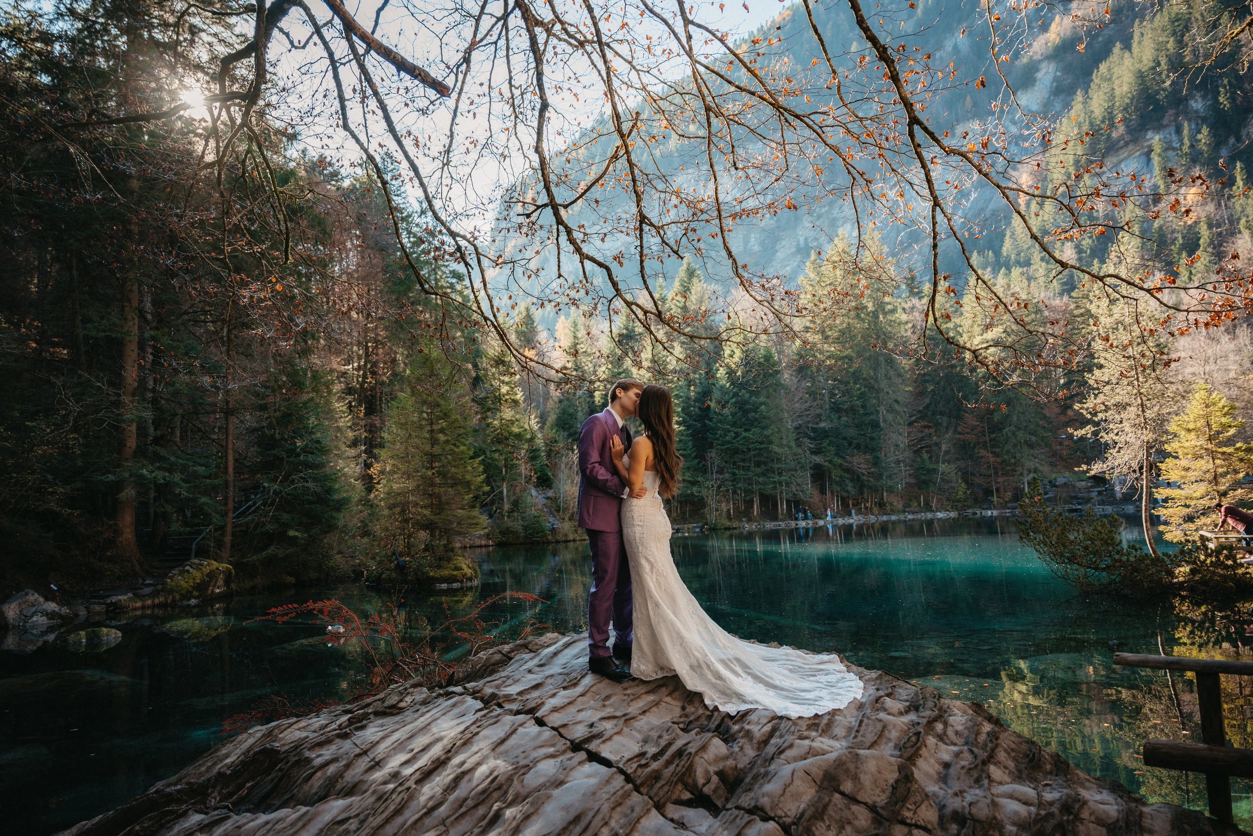 Pareja en vestido de novia y traje en un entorno natural junto a un lago rodeado de árboles en un paisaje montañoso.