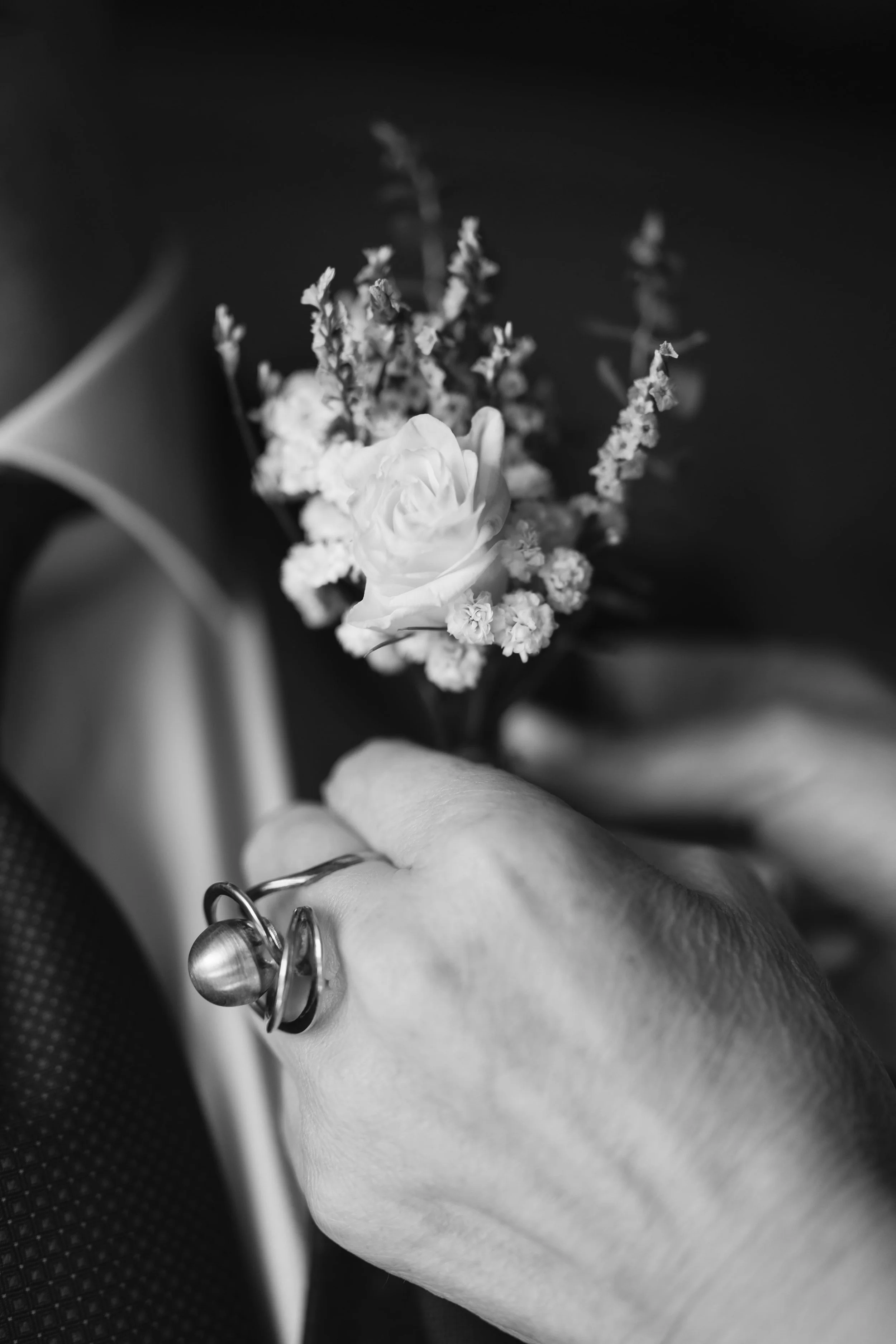 Mano con anillos sosteniendo un pequeño ramillete de flores blancas, en una fotografía en blanco y negro.