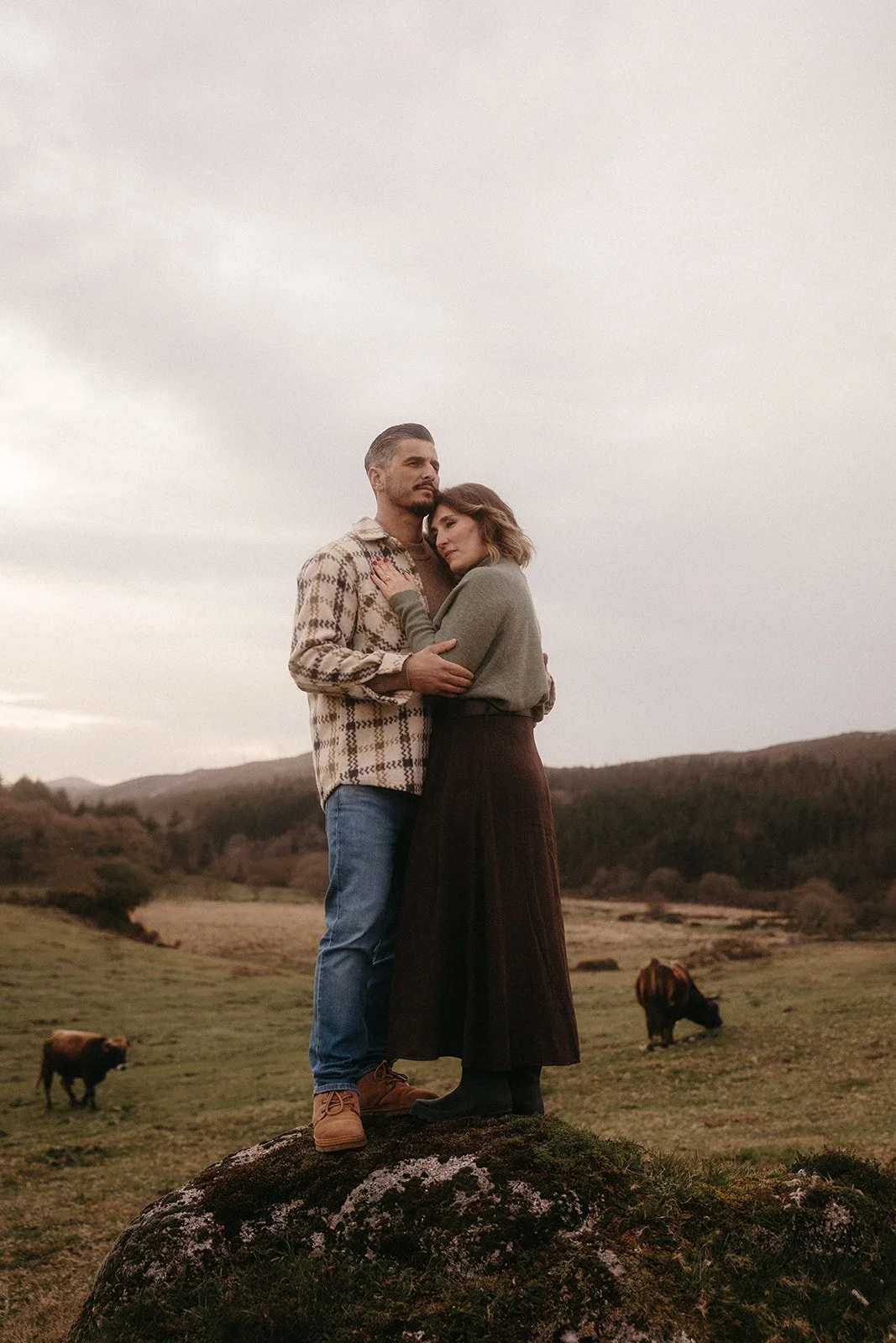 Una pareja abrazada en un paisaje de campo con montañas de fondo, junto a dos bisontes.