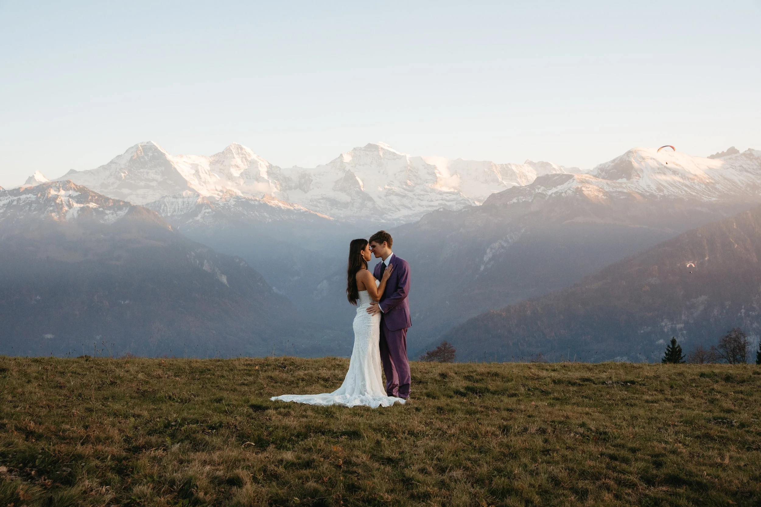 Pareja de novios en vestido de boda y traje en un campo con montañas nevadas de fondo.