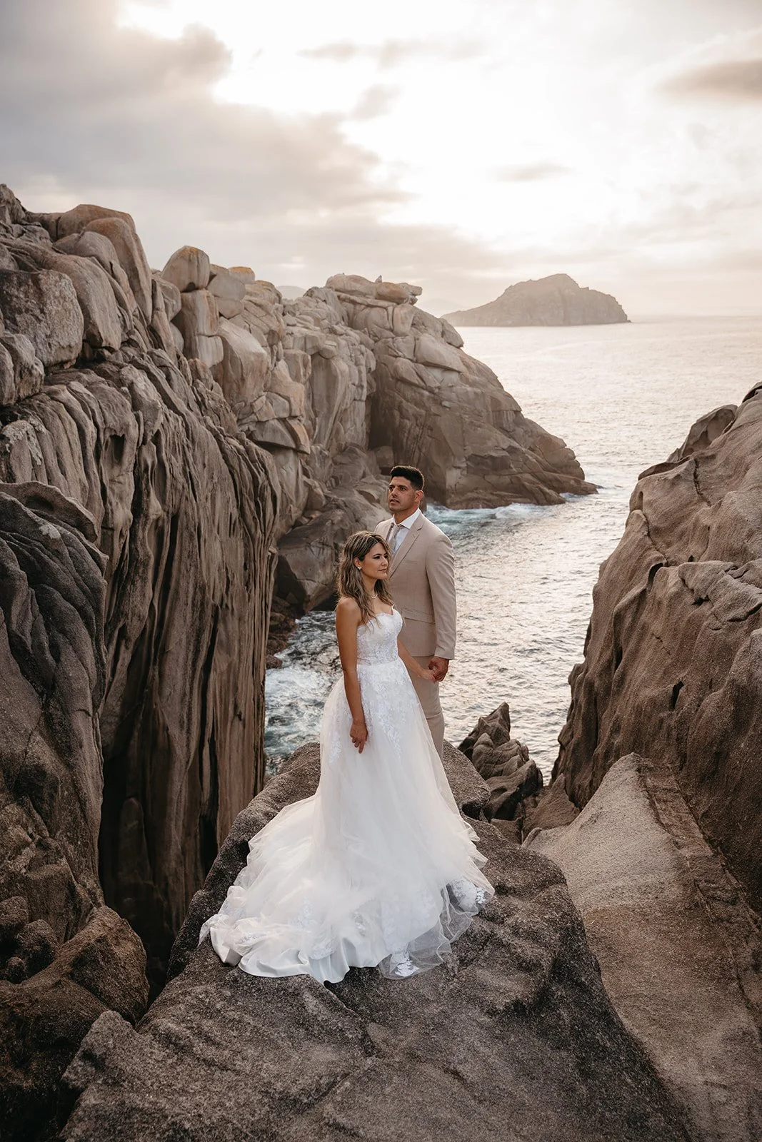 Una pareja en ropa de boda en un paisaje costero con rocas y el mar al fondo, durante el atardecer en un lugar aislado y rocoso.
