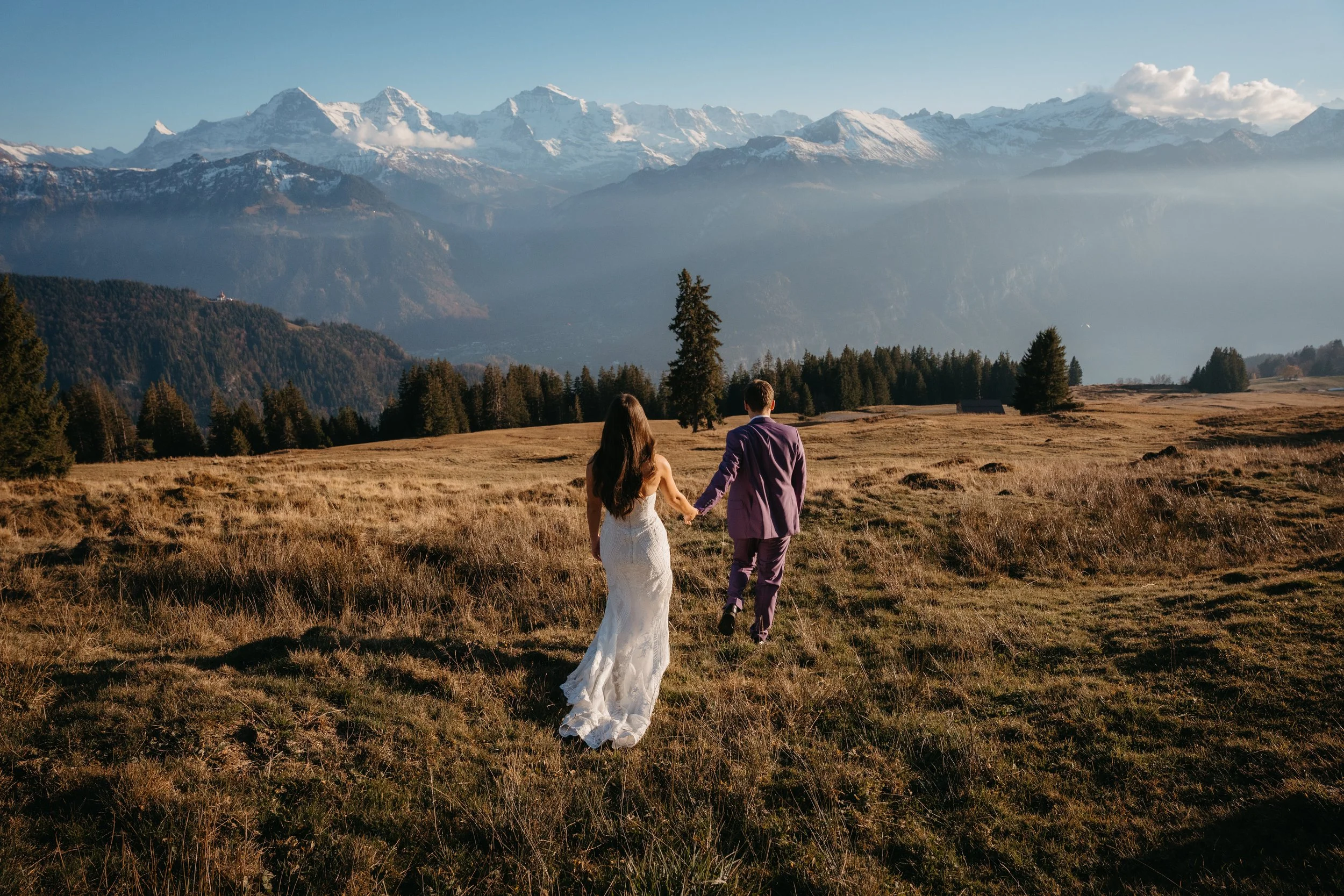 Una pareja vestida de novia y novio caminando por un prado con montañas nevadas al fondo, en un paisaje natural y hermoso.