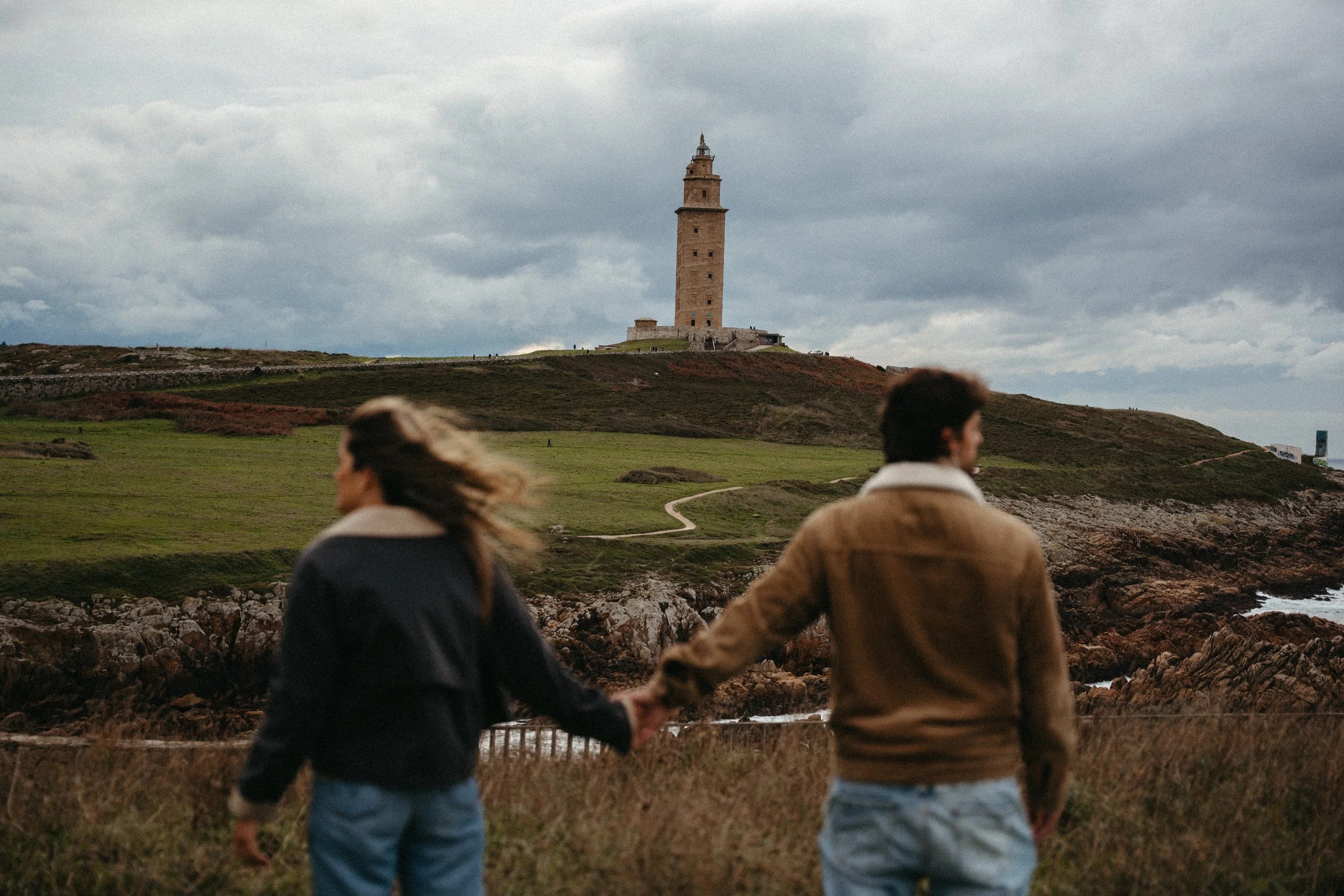Una pareja de jóvenes sostenidos de la mano, de espaldas, en primer plano. En el fondo, la Torre de Pisa sobre una colina y un cielo nublado.