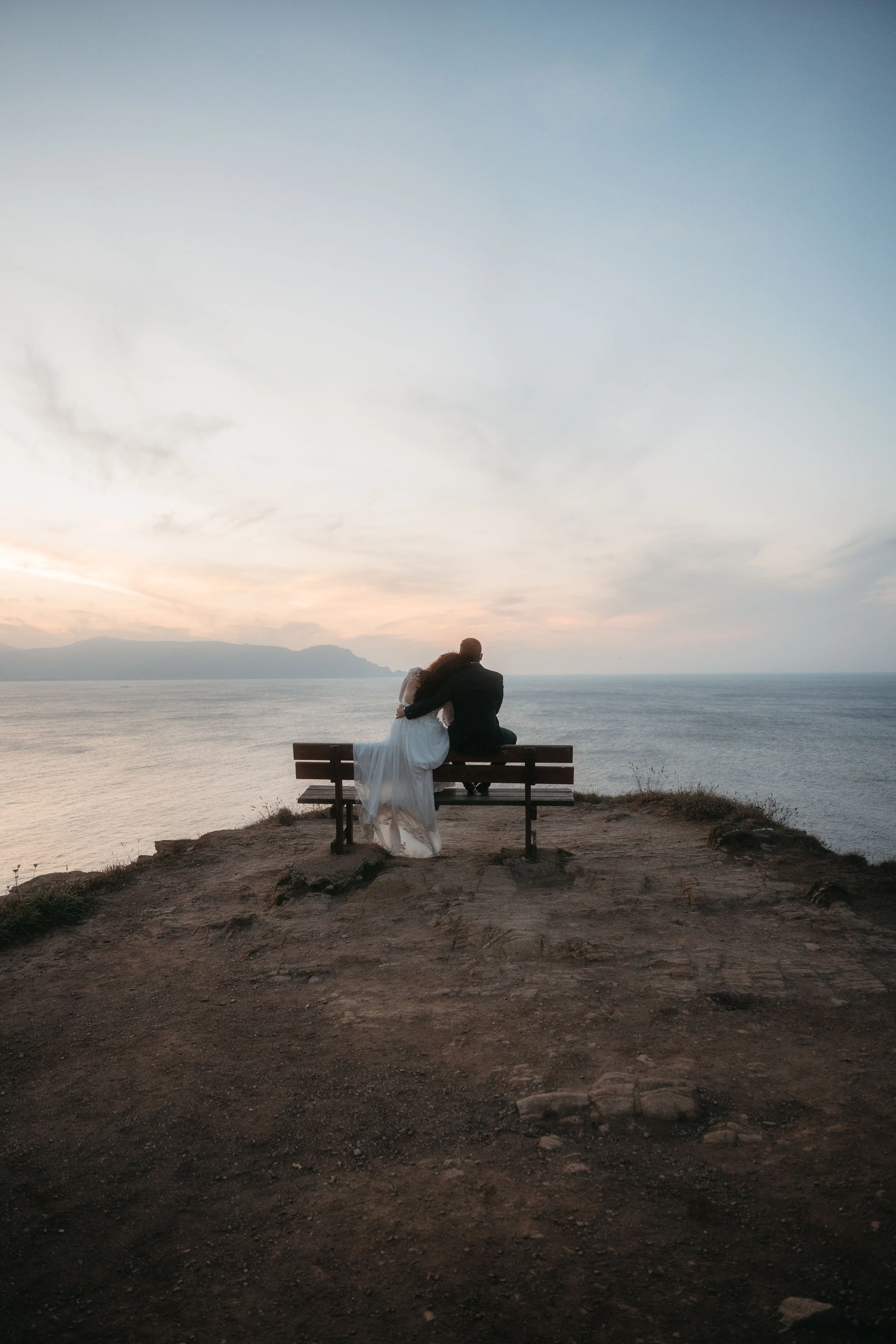Una pareja sentada en un banco en un acantilado mirando el mar durante el atardecer.