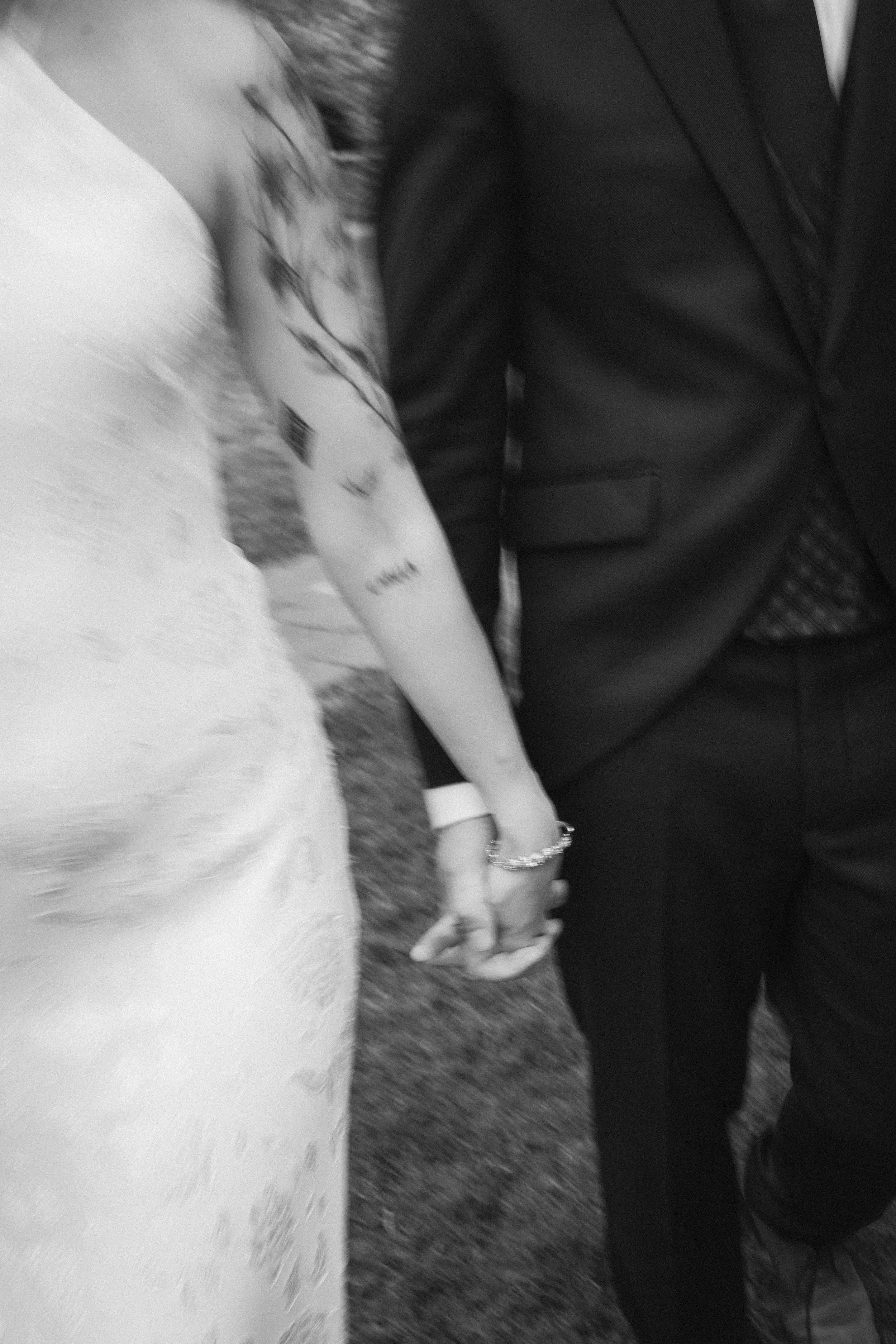 Una pareja de novios tomados de la mano en su boda, ella con vestido blanco y él con saco negro.