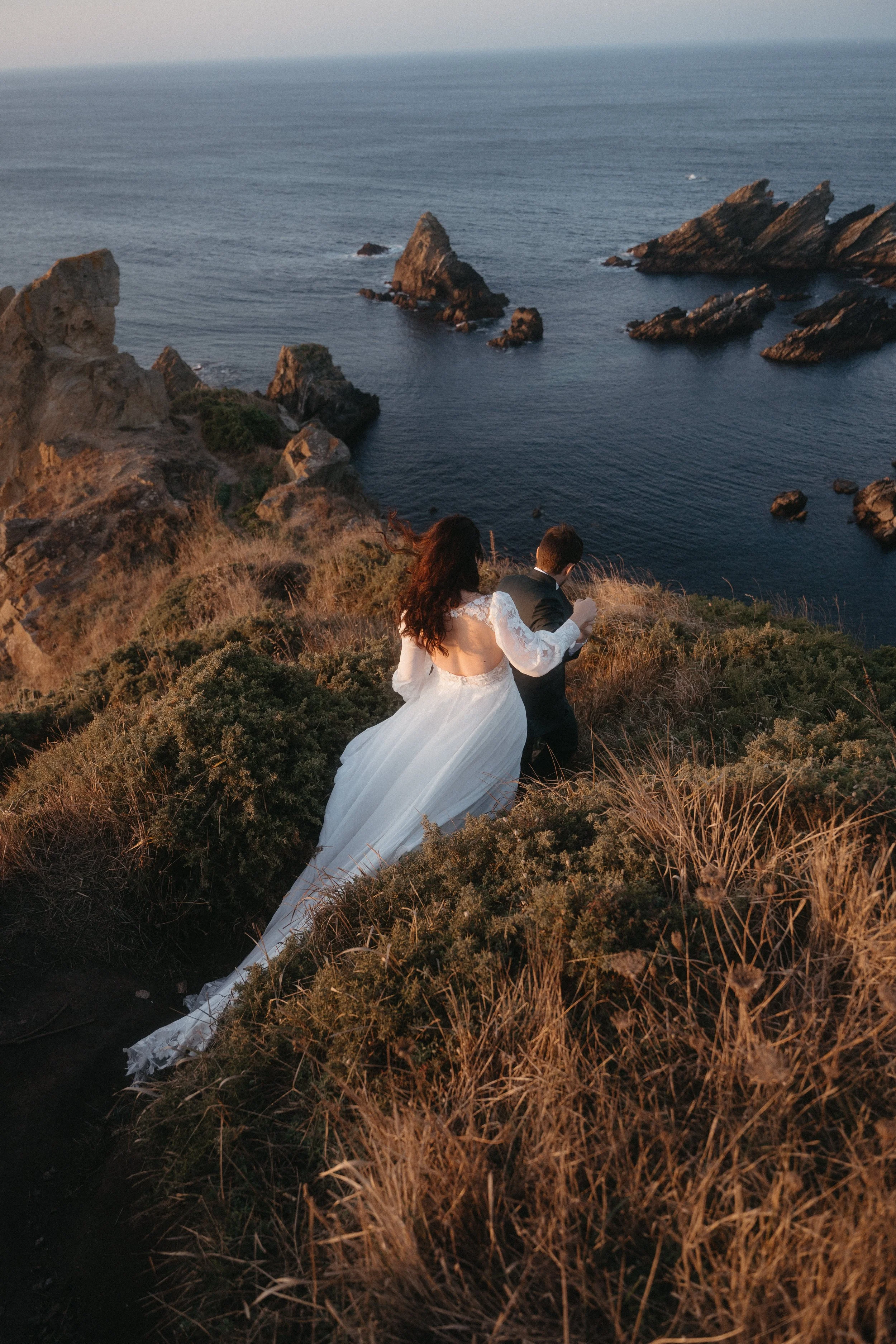 Una pareja de novios camina por un acantilado con vista al océano, en el atardecer, con rocas en el mar.