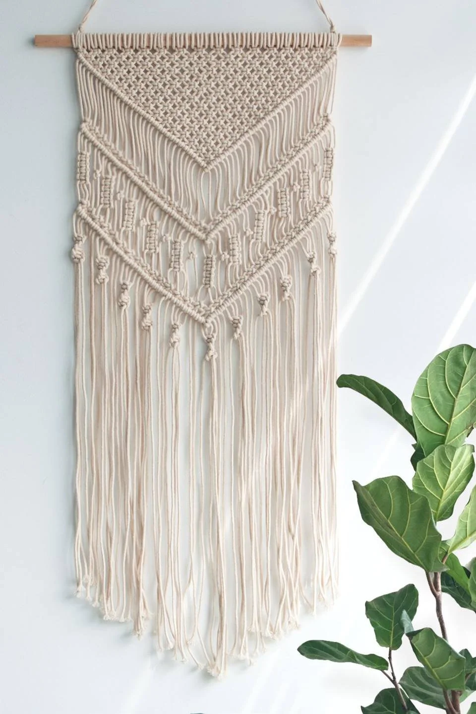 A wall hanging made of cream-colored macramé with a wooden rod at the top, featuring intricate patterns and long fringes at the bottom, hanging on a light blue wall near a green leafy plant.