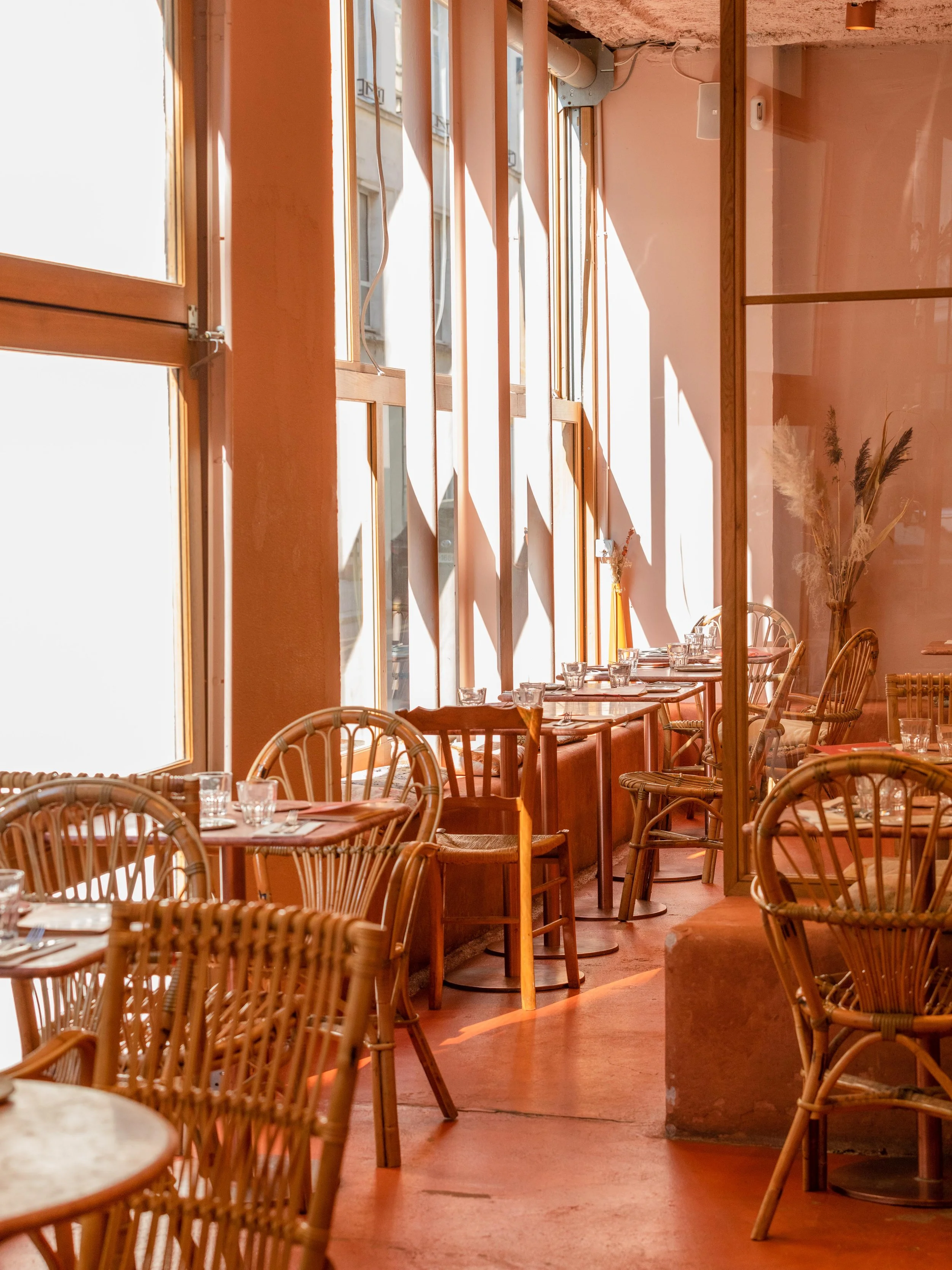 Salon de restaurant lumineux avec des tables en bois, chaises en rotin, et de grandes fenêtres laissant entrer la lumière naturelle.