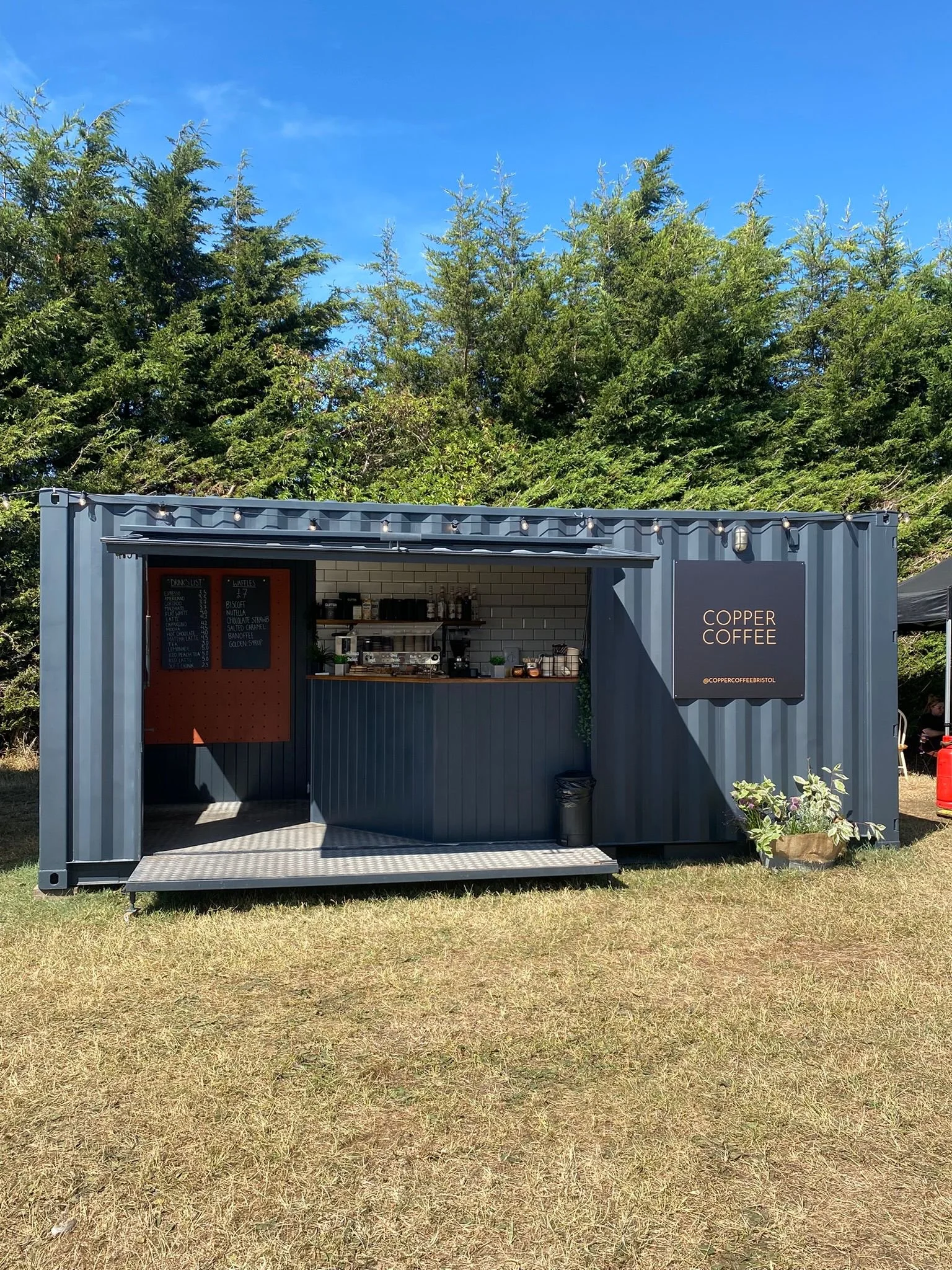 a dark grey shopping container with Copper Coffee sign on the front large opening in the front to a coffee bar inside serves good coffee in a sunny field