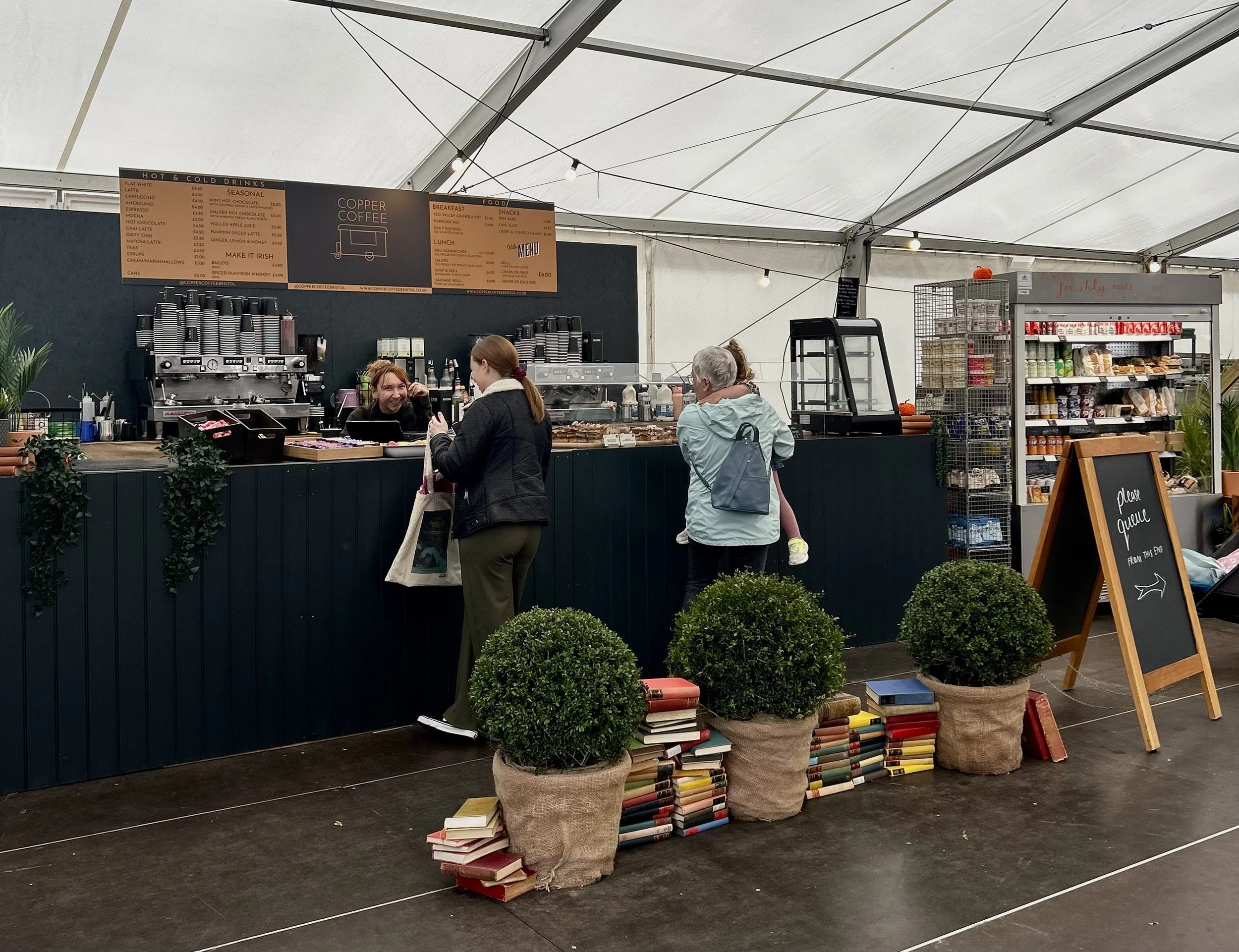 customers are served by a happy barista at our customisable bar cafe with anthracite grey cladding, two La Marzocco coffee machines, multi-deck fridge display, cakes an large menu boars on the walls