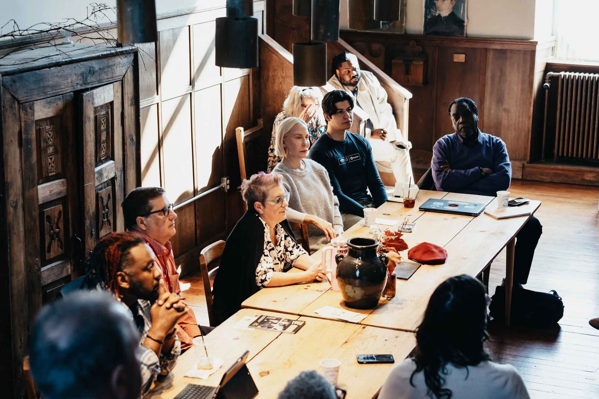 A diverse group of people sitting around a large wooden table in a meeting room, listening attentively with laptops, notebooks, and drinks in front of them.
