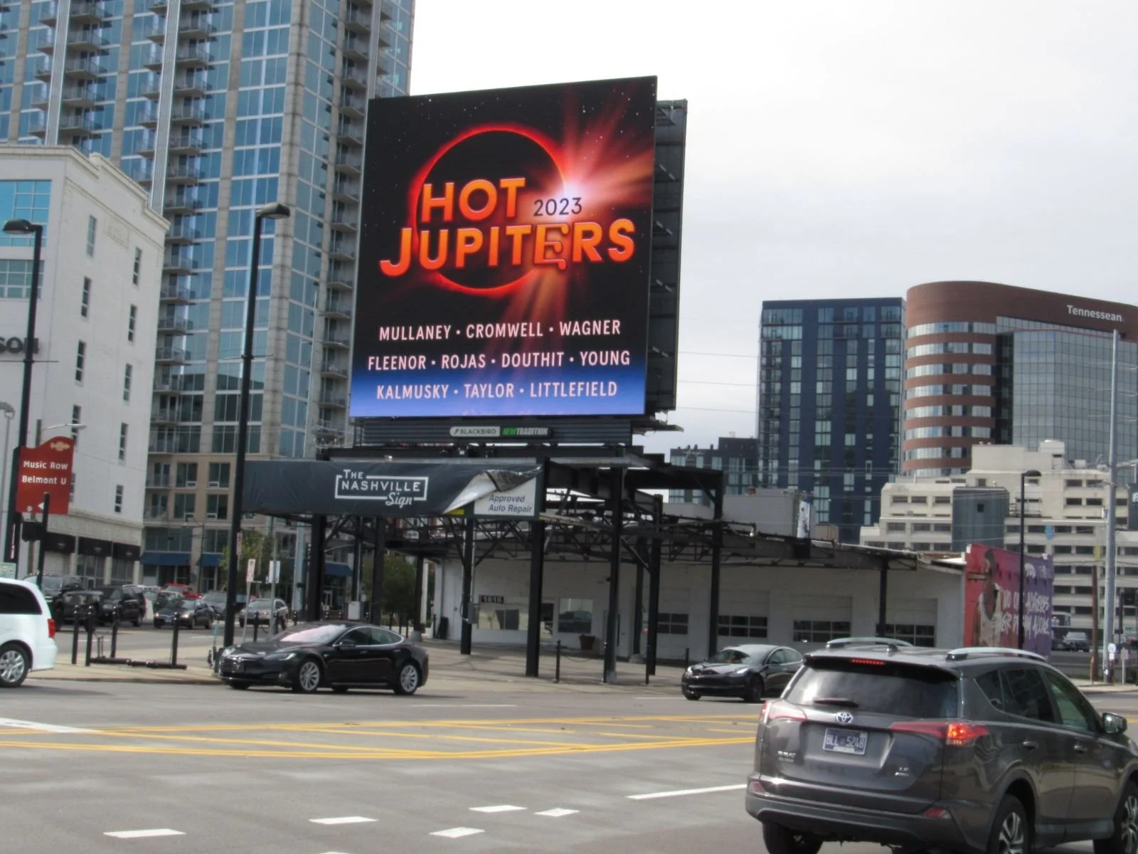 Digital billboard advertising the 2023 event "Hot Jupiters" with a list of performers, set against a cityscape with tall buildings and cars on the road.