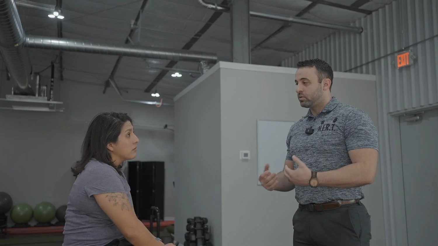 A woman sitting on a workout bench listening to a male fitness trainer explaining exercises in a gym.