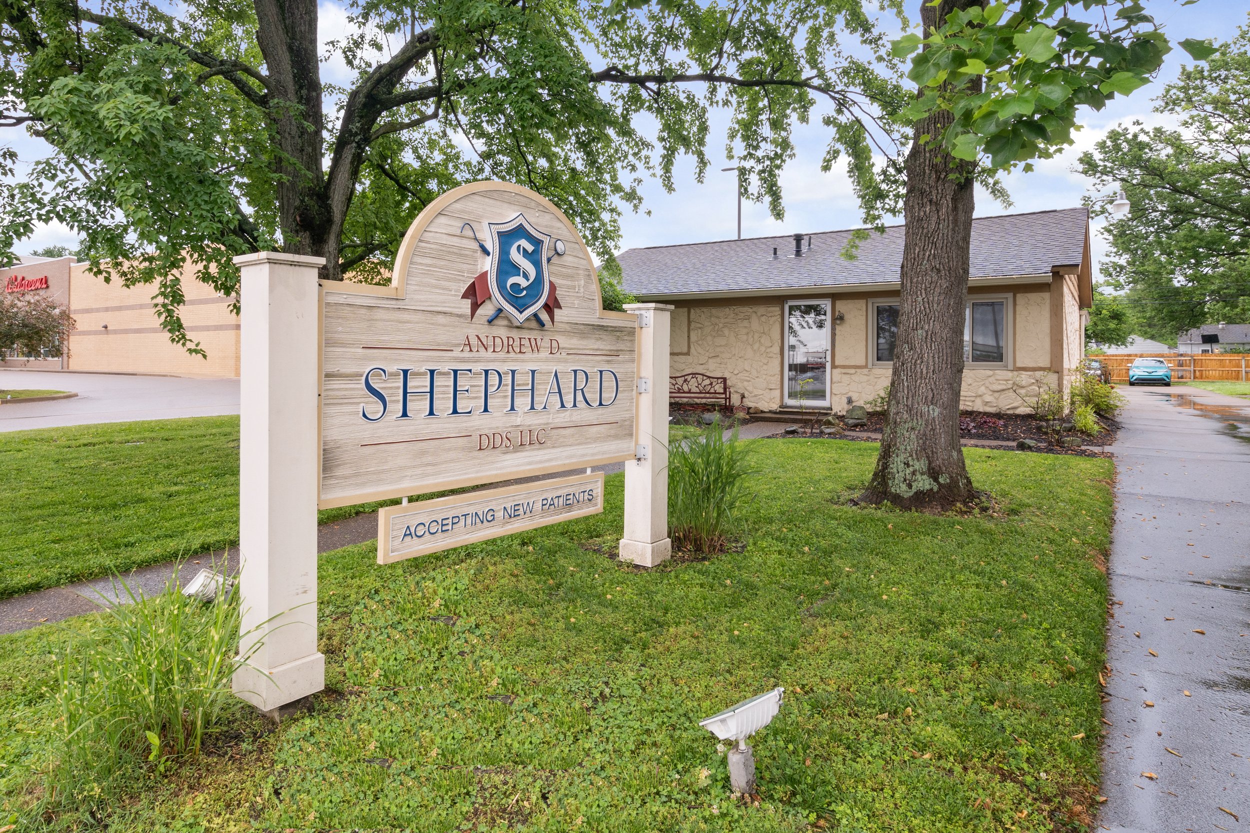 Wooden sign reading: "Andrew D. Shephard DDS Inc. – Accepting New Patients" in front of a stone dental office building surrounded by trees and green lawn.