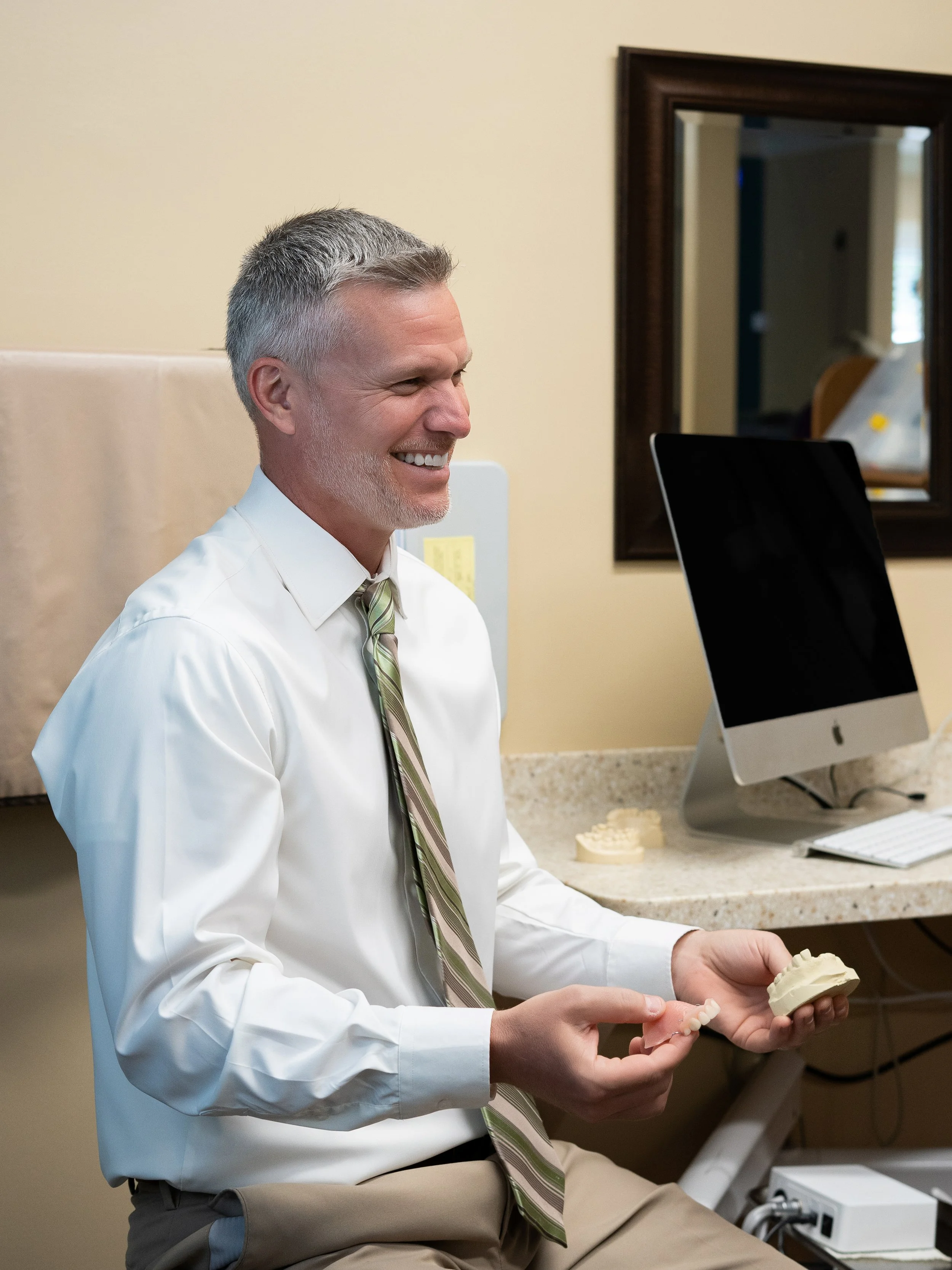 Dentist in a white dress shirt and tie smiling while holding a dental model in a treatment room.