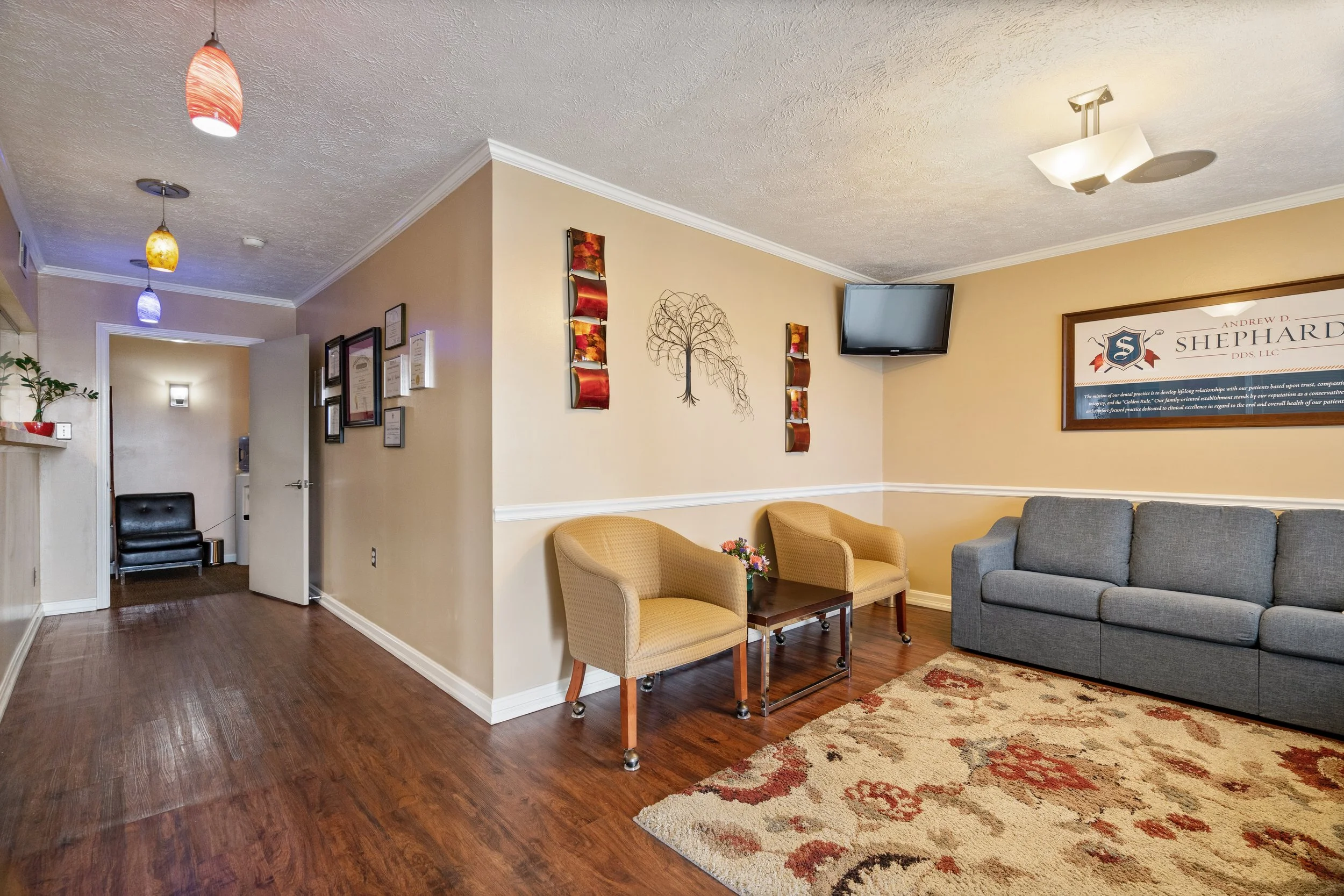 Cozy dental office waiting room with hardwood floors, warm beige walls, chairs, a gray sofa, patterned rug, wall-mounted TV, and framed artwork.