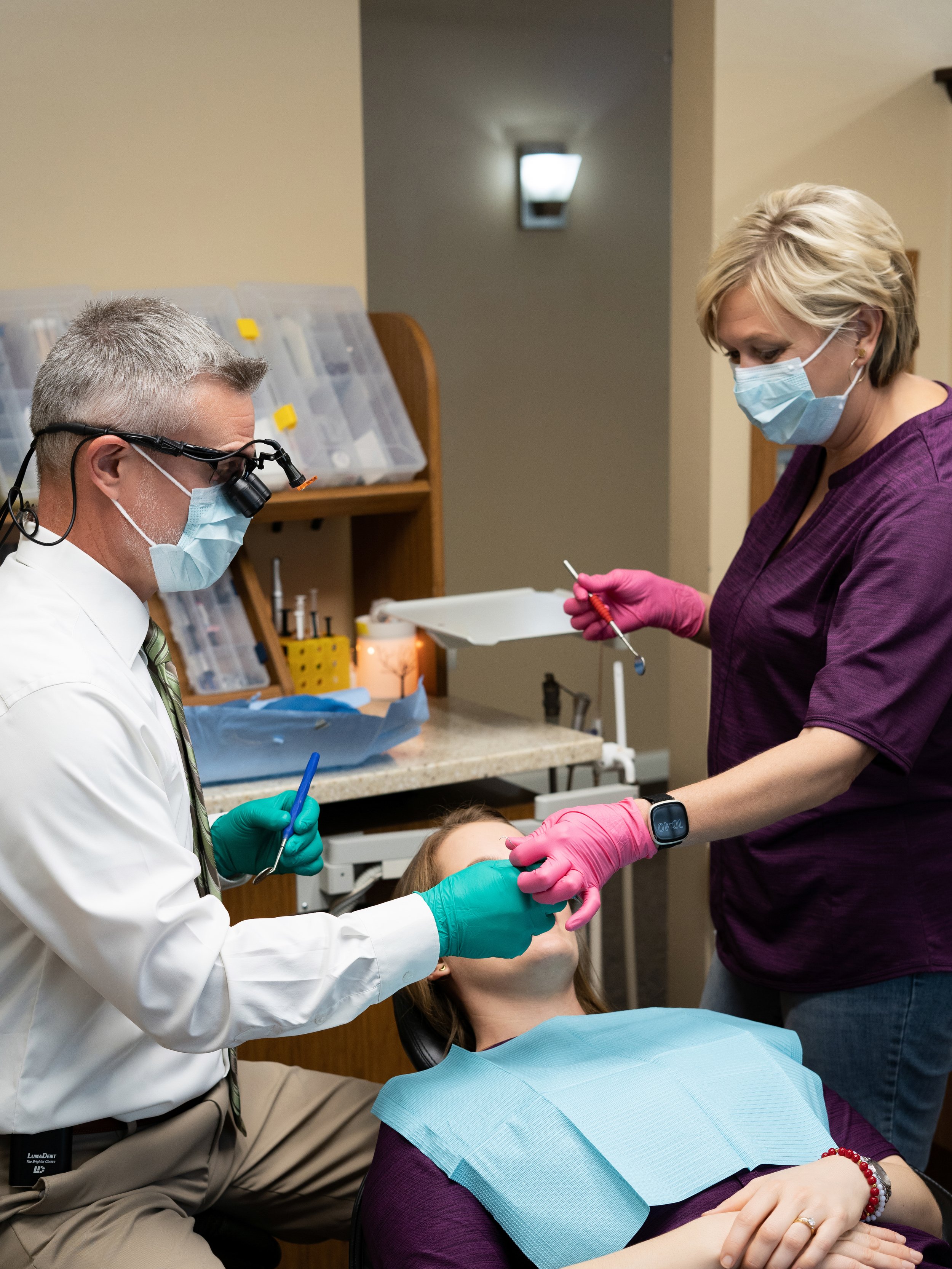 Dentist wearing loupes and a mask with green gloves working alongside a dental assistant in pink gloves who is holding instruments, with a patient seated in the treatment chair between them.