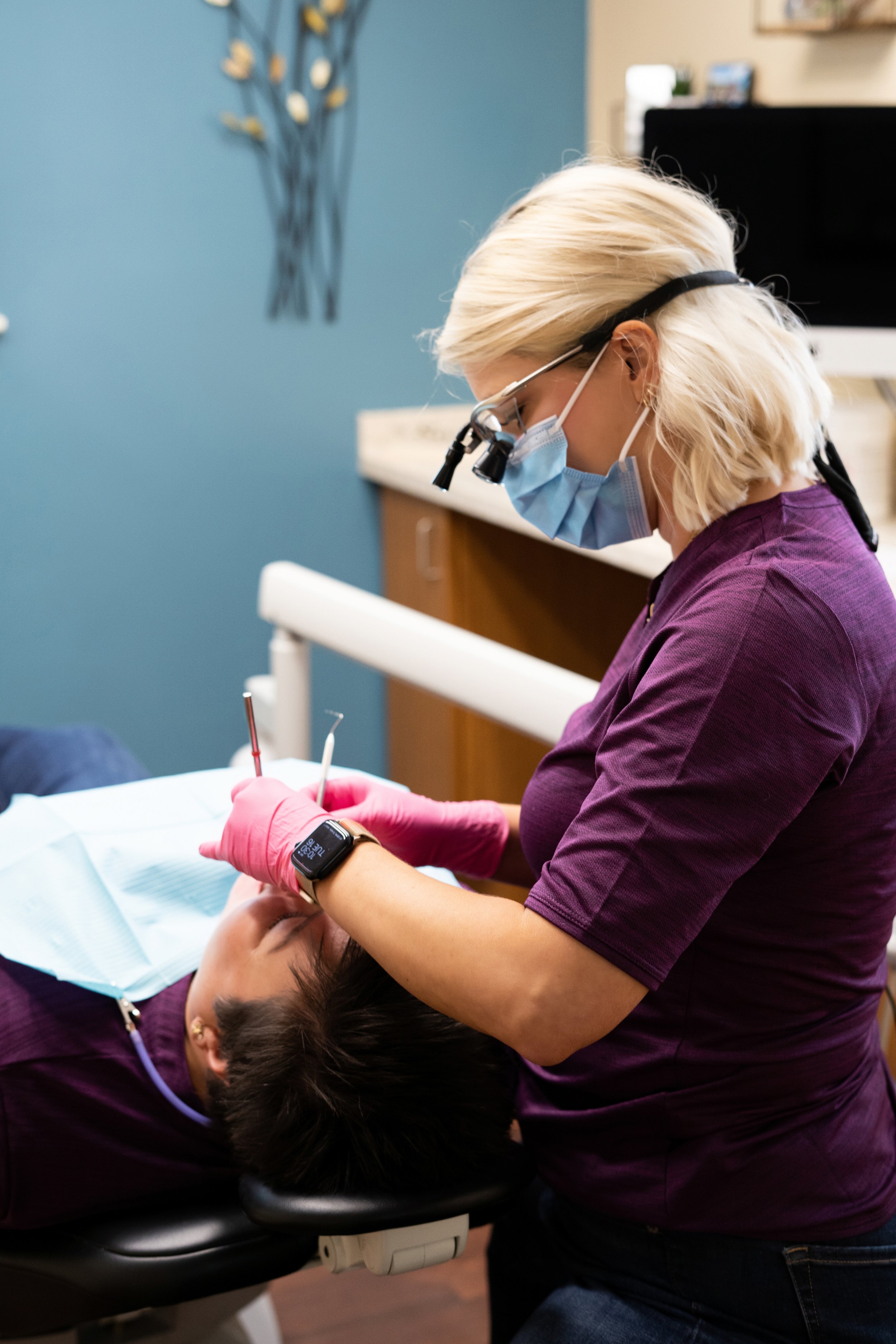 Dental hygienist wearing magnifying loupes, a blue mask, and pink gloves performing a dental cleaning on a patient reclined in a dental chair.