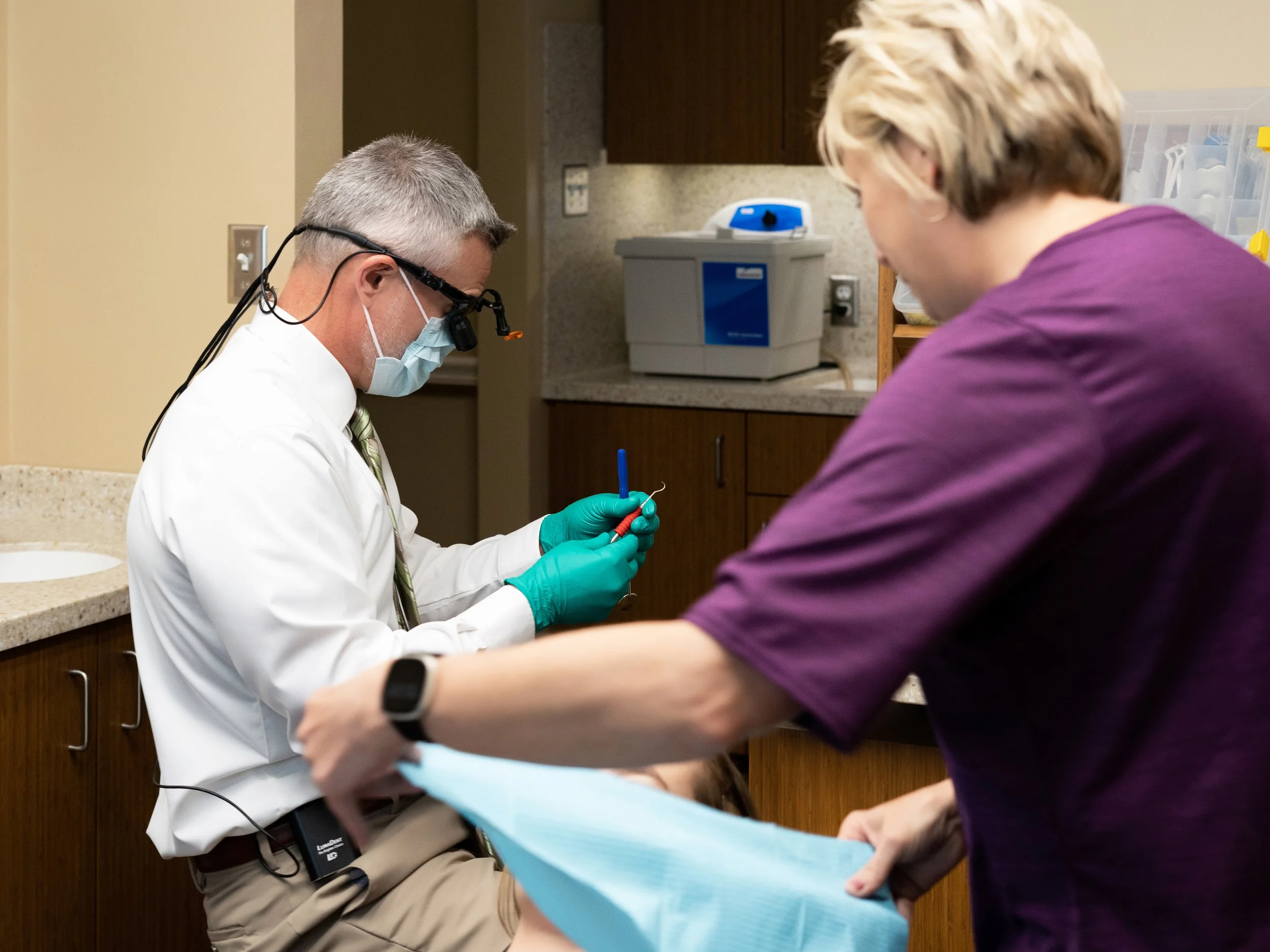 Dentist wearing magnifying loupes, surgical mask, and green gloves preparing dental instruments while a dental assistant in a purple top arranges a patient bib in a treatment room.