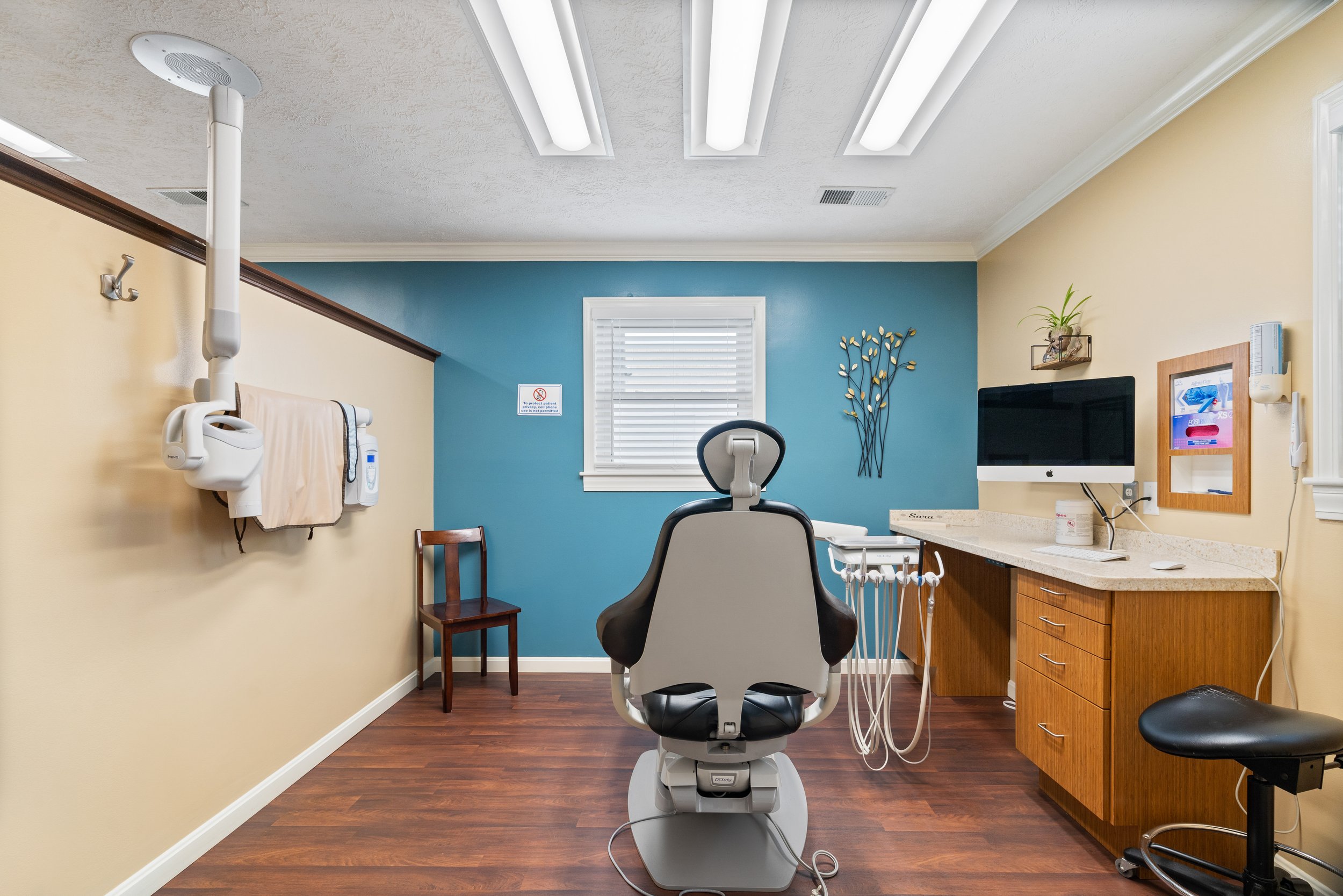 A clean, modern dental treatment room with hardwood floors, a teal accent wall, dental chair, wall-mounted X-Ray equipment, and a computer workstation.