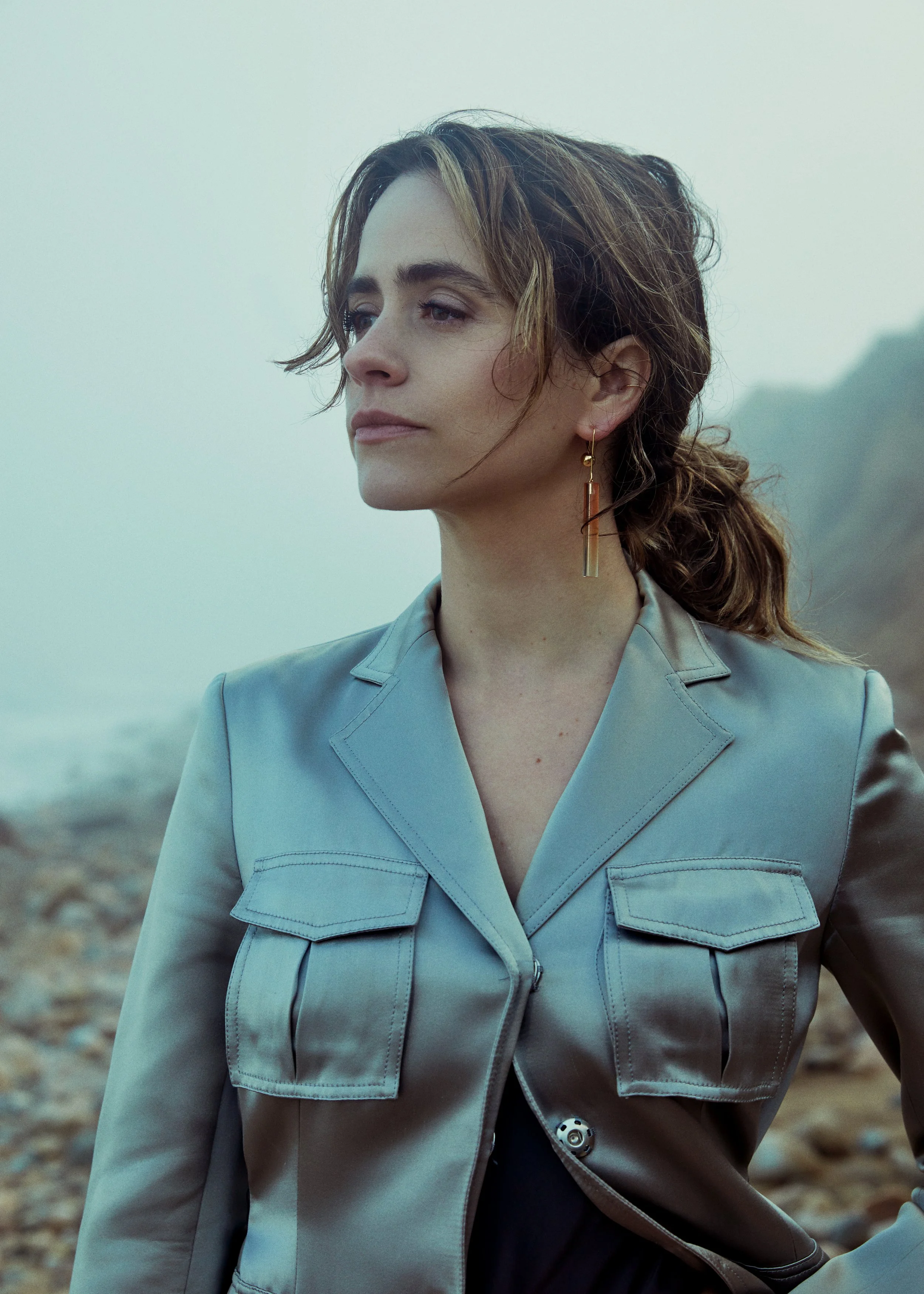 A woman with wavy brown hair tied in a loose ponytail, standing outdoors on a rocky coastline with foggy cliffs in the background, wearing a light blue jacket and dangling earrings.